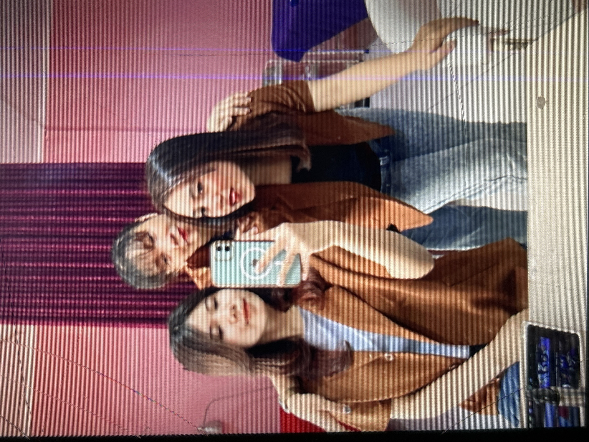 The image depicts a mirror selfie taken in what appears to be a beauty salon or hair salon in Depok, Indonesia. Three young women are visible. The woman on the left is holding her phone to take the selfie, and her left arm is around the shoulder of the woman in the center. The woman in the center is leaning in slightly. The woman on the right has her arm around the woman in the center's shoulder from the other side.
All three women have dark hair and appear to be wearing makeup. They are all dressed casually. The woman on the left wears a brown blazer over a light blue top. The woman in the center wears a brown blazer over a black top. The woman on the right wears a brown blazer over a black top and blue jeans.
The background features a pink wall with horizontal lines, and a section of dark purple curtains. There are also elements of salon equipment visible, including a white chair on the right and a metal cart behind the woman on the right. The overall lighting suggests an indoor setting. The image is slightly distorted due to a screen overlay with horizontal lines.