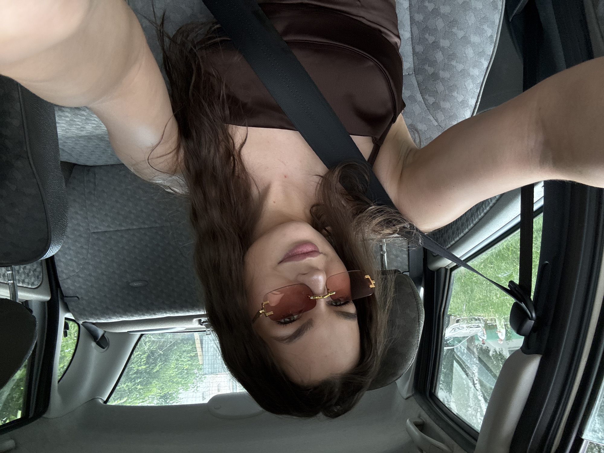 A young woman with long, dark brown hair and wearing sunglasses and a dark brown top is taking a selfie inside a car. She is sitting in the driver's seat and her left arm is extended towards the camera. The car's interior is visible, including the gray patterned seats and a black seatbelt. Outside the car window, a street scene is visible with greenery, trees, and two men standing near a silver car. This image appears to have been taken in the city of Chaikovsky, Russia.