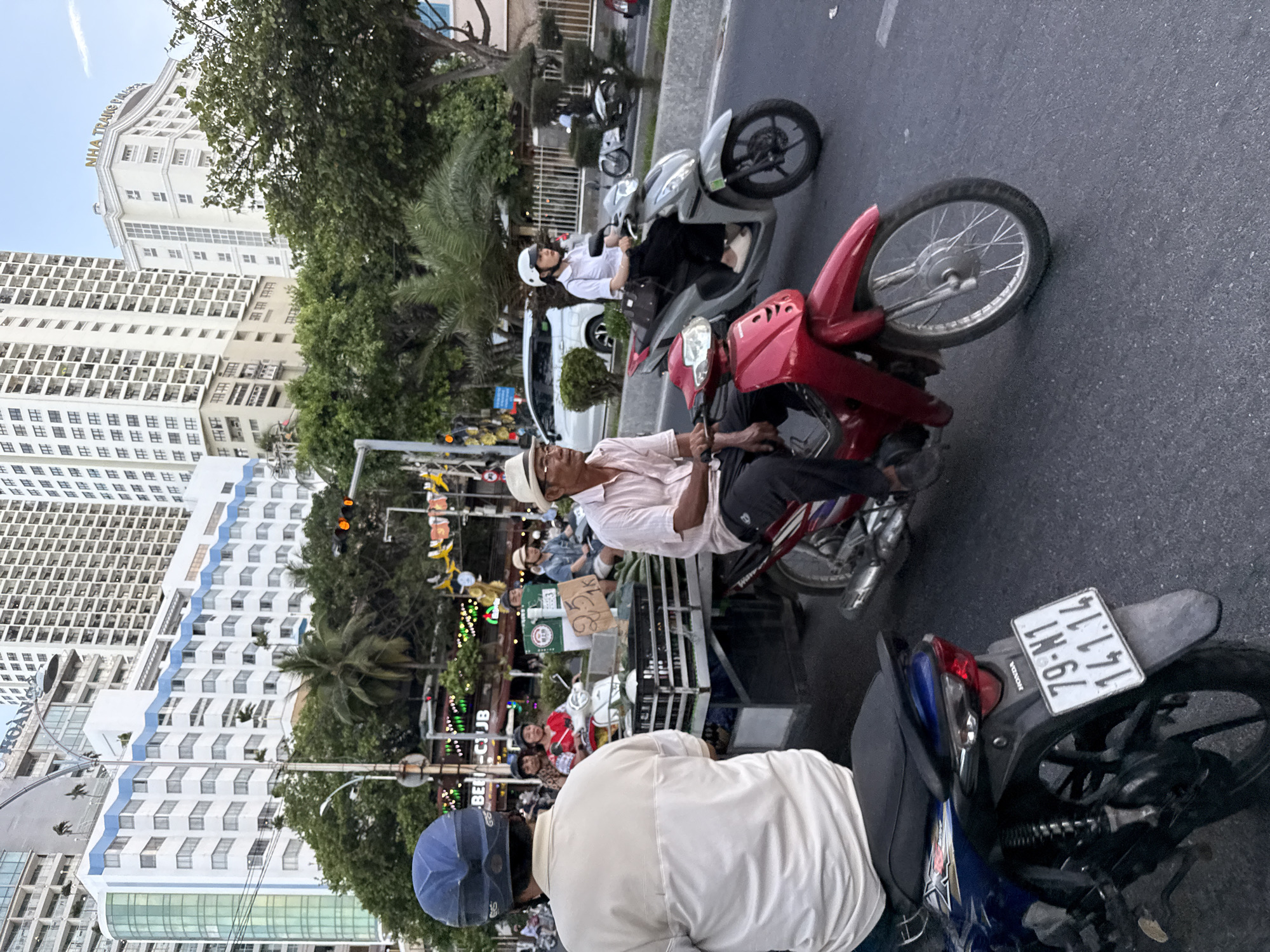 This media file captures a dynamic street scene in Nha Trang, Vietnam, likely during the daytime given the bright lighting and shadows. The image is oriented vertically, showing a bustling street with several people on motorcycles.
In the foreground, two individuals are prominently featured. The person on the left, wearing a white shirt and a straw hat, is partially visible from behind. To their right, another person in a white shirt and helmet is riding a red motorcycle. Behind them, a person in a white shirt and helmet is on a silver motorcycle. Further back, a man in a white shirt and dark pants is also on a motorcycle.
The setting includes modern buildings with multiple windows, suggesting an urban environment. Lush green trees and palm trees are interspersed with the architecture, adding a tropical feel. A variety of motorcycles, some with visible license plates, are parked or in motion. One license plate clearly reads "79-N1 141.14". Signs with text such as "HOÀNG" and "BEE-CJB" are visible, hinting at businesses or street names. Decorative lights are also present, suggesting a festive or commercial area.
The overall atmosphere appears to be that of daily life on a busy street, with people commuting or engaged in commercial activities. The orientation and angle of the photo provide a unique perspective, looking down and across the street.