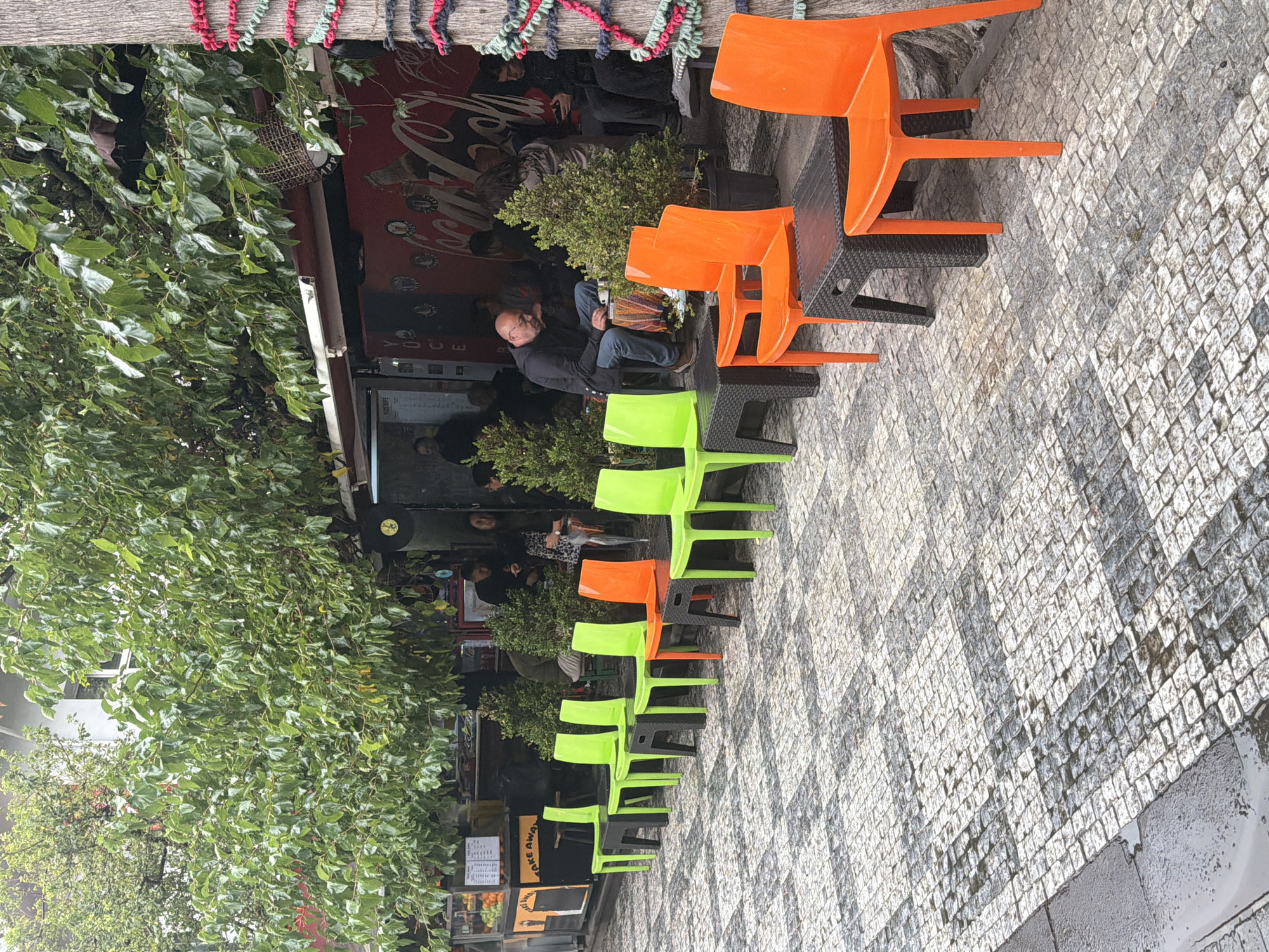 The image depicts an outdoor seating area in front of a building in Kadıköy, Turkey. The seating area is paved with cobblestones that appear wet, suggesting recent rain. A row of brightly colored plastic chairs, predominantly green and orange, are set up with small, dark-colored tables. Some of these chairs are occupied by people.
In the background, there is a building with an awning and a red facade featuring Coca-Cola branding. Several individuals are seated at tables or standing near the building entrance. A man in a black jacket is seated at a table, looking down at something in his lap. To his right, other people are visible, some with their phones. Above the seating area, lush green foliage hangs from trees, providing shade.
To the left of the seating area, a food stall or kiosk is visible with displayed fruits and a "TAKE AWAY" sign. The overall impression is of a casual, outdoor cafe or eatery on a street in Kadıköy.