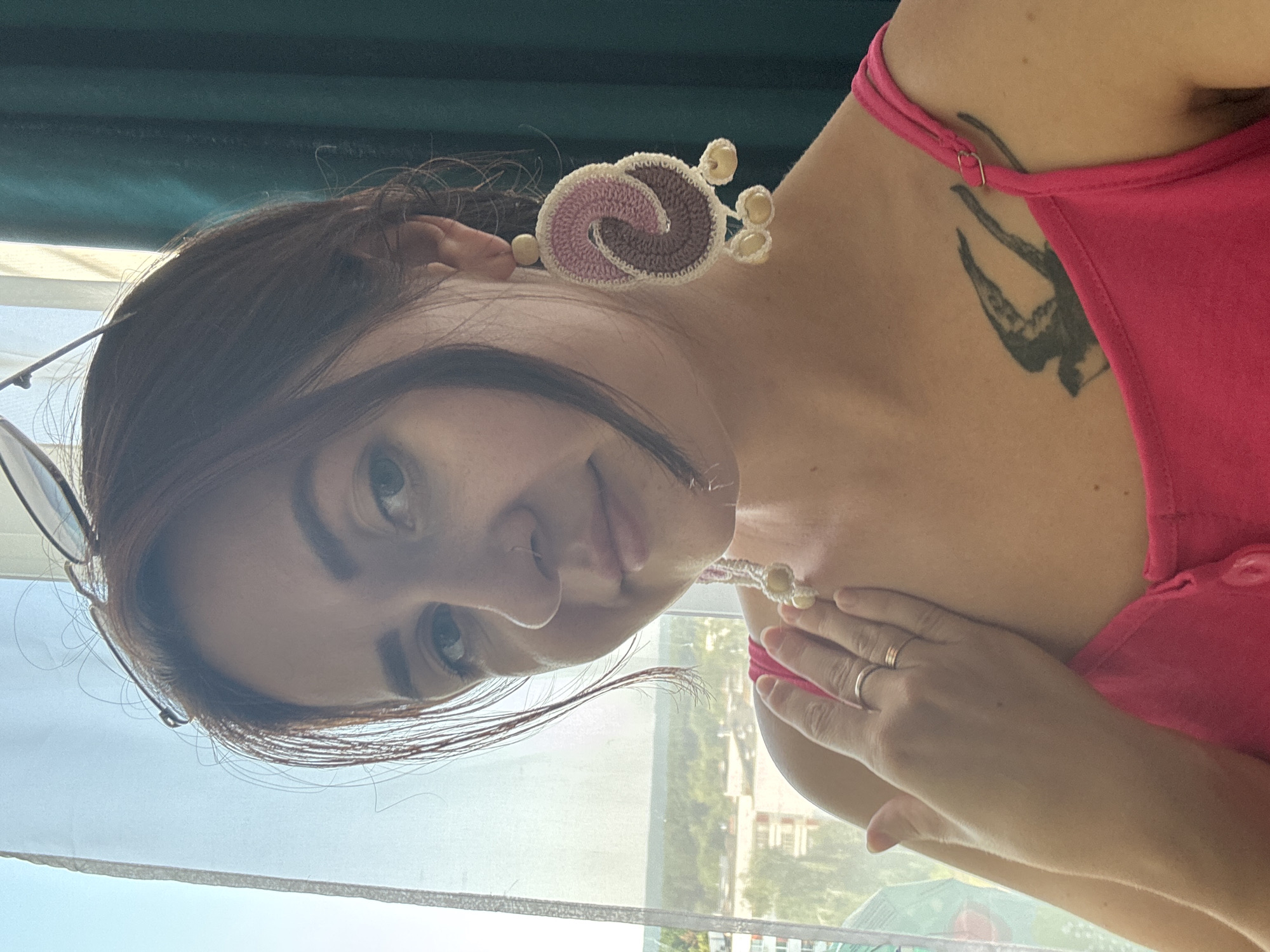 The image shows a young woman with brown hair and light blue eyes, wearing a pink tank top. She has a tattoo of a bird on her chest and is wearing large, crocheted earrings with a pink and brown swirling design. She is also wearing a silver ring on her left hand, which is placed on her shoulder. The woman is indoors, with a window and curtains visible in the background. Outside the window, there is a view of buildings and trees, suggesting an urban or suburban setting. The lighting suggests it is daytime. The photographer is close to the subject, capturing a close-up of her upper body and face. Based on the presence of a prominent billboard in the background featuring text in the Russian language and Cyrillic alphabet, it is likely that this photo was taken in Russia, possibly in the city of Naberezhnye Chelny, given its known industrial and urban landscape.