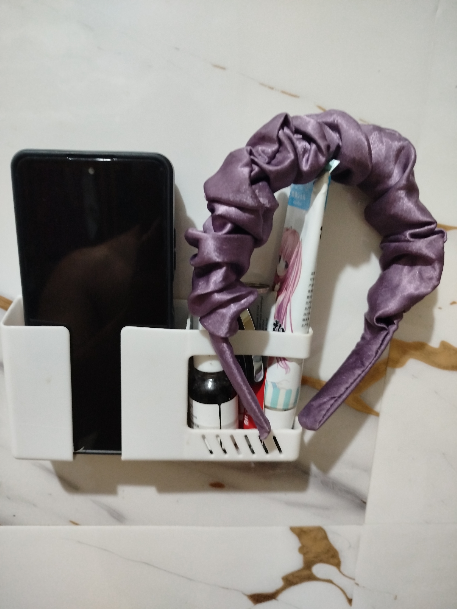 In Bandungrejo, Indonesia, a white wall-mounted organizer holds a black smartphone in its left compartment. The right compartment contains a tube of what appears to be toothpaste, a small dark bottle with a white label, and what looks like another tube or roller. A scrunchie made of shiny purple fabric is draped over the organizer, with one end resting inside the right compartment and the other extending outwards. The wall behind the organizer has a marble-like pattern with brown and grey veins.