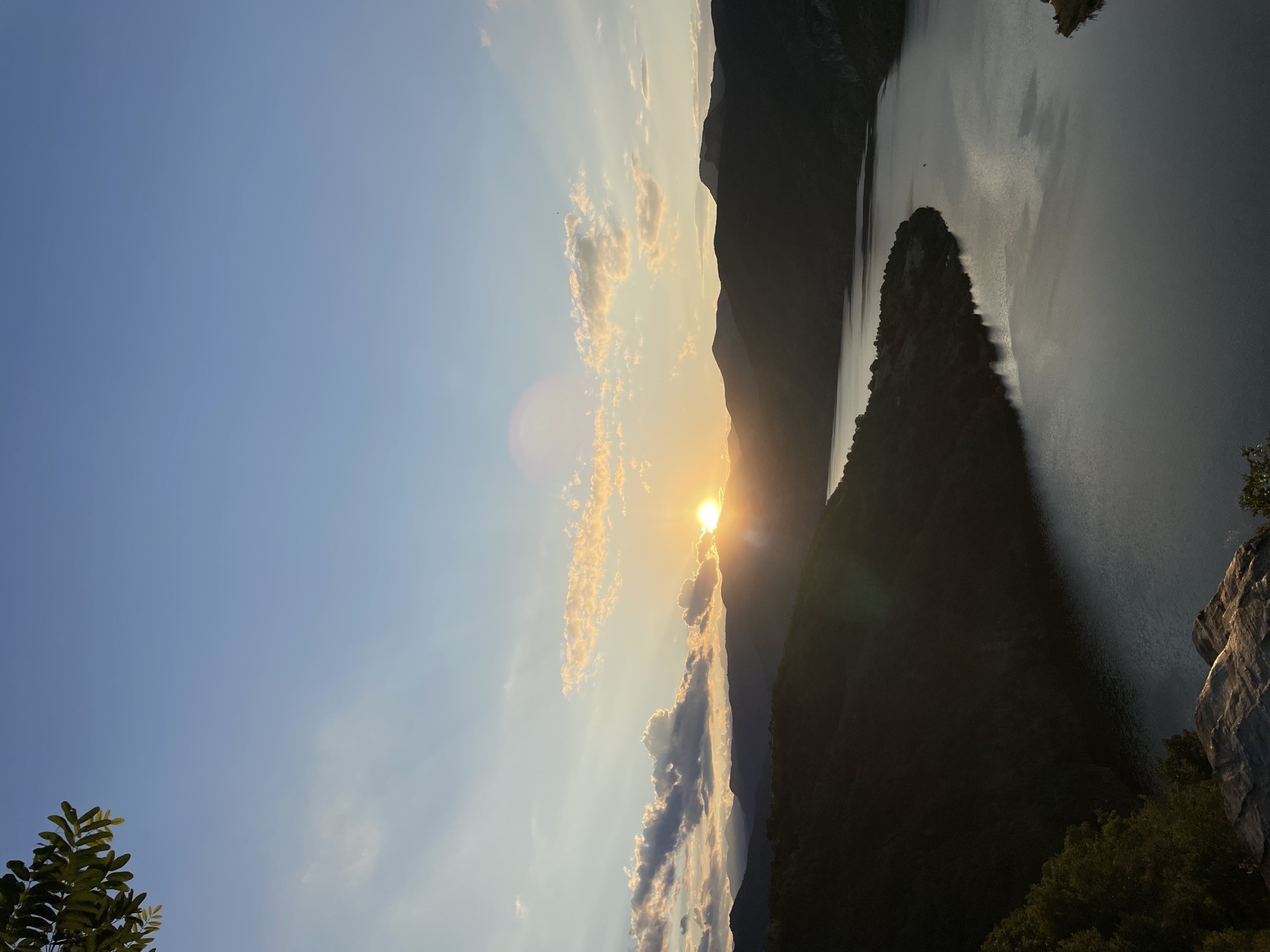 A high-angle, vertical outdoor shot captures a serene sunset over a winding river flanked by dark, tree-covered mountains. The sun, a bright orb tinged with orange and yellow, is partially obscured by wispy clouds and rays of light pierce the sky, casting a warm glow. The river, a dark, reflective surface, mirrors the sky and mountains, its surface disturbed by gentle ripples. In the foreground, a rocky ledge offers a vantage point, with a few green leaves visible in the upper left corner. The scene is devoid of human presence, creating a sense of tranquil solitude. The image evokes the natural beauty of the location in Borovitsa, Bulgaria, during twilight hours.