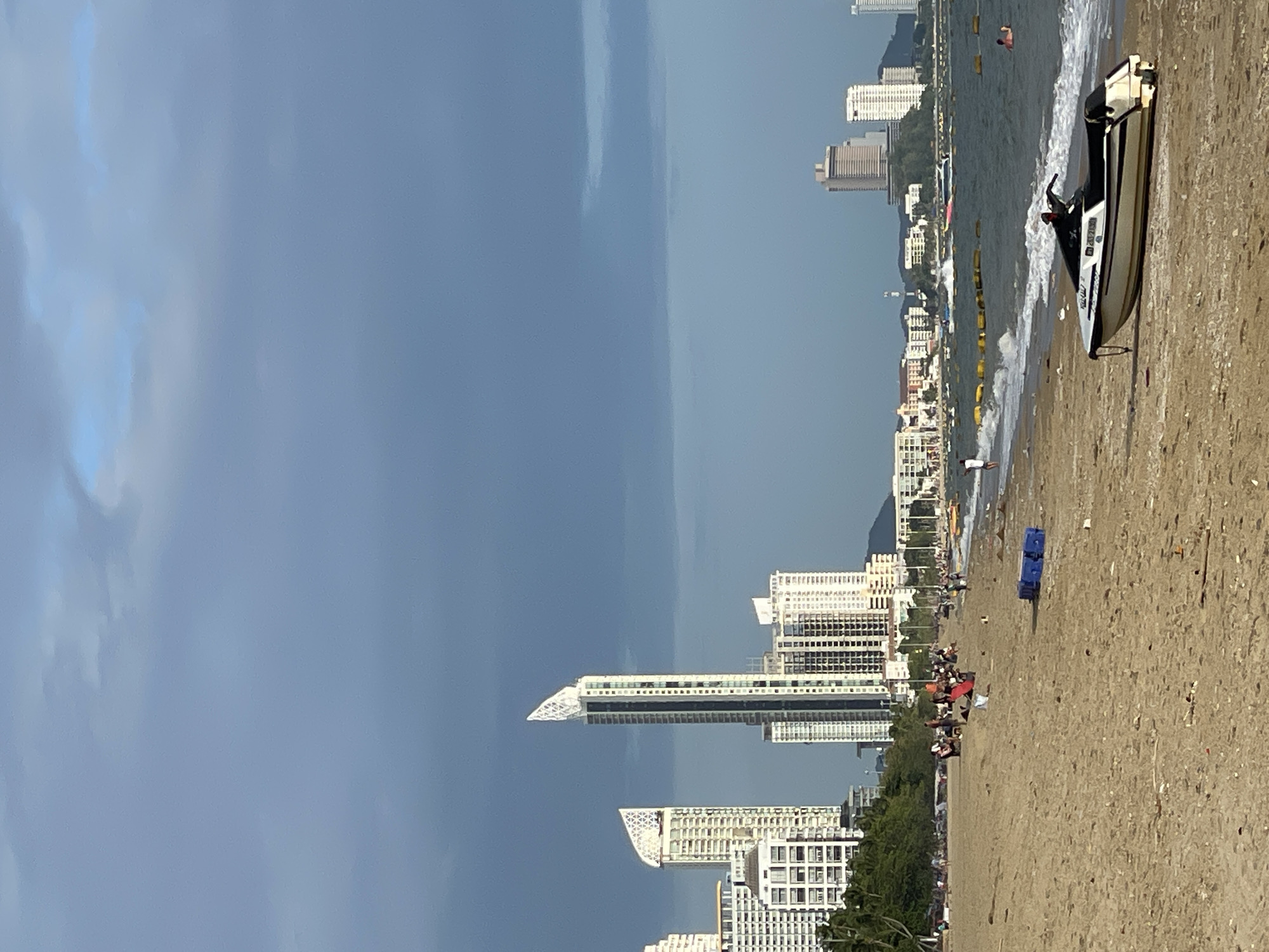 This is a full shot of a beach in Pattaya City, Thailand, under a cloudy sky. The foreground is dominated by a sandy beach with scattered debris. A white and black jet ski is parked on the sand near the water's edge. In the middle ground, people are scattered across the beach, some sitting and others walking. The water is choppy with waves breaking on the shore. A blue plastic container is visible on the sand. In the background, a cityscape of tall buildings and hotels lines the coast, with lush green trees interspersed. A few boats are visible on the water in the distance. The overall atmosphere is that of a late afternoon at a busy beach destination.