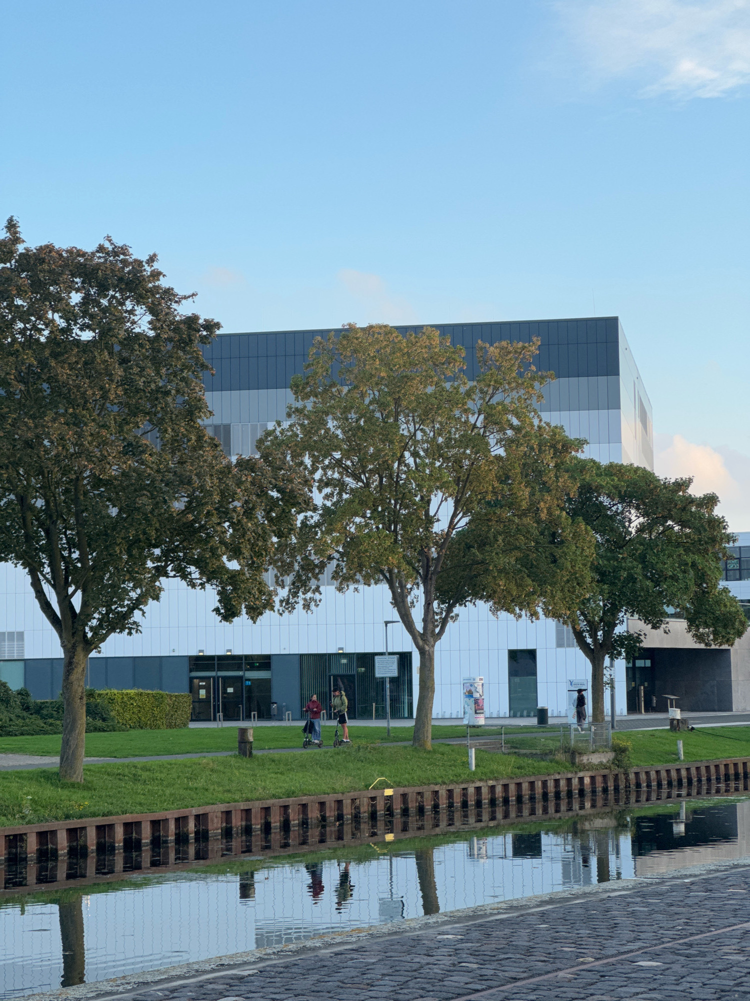 In Cleves, Germany, a modern building with white and dark grey panels faces a calm canal. Three trees stand between the building and the water. On the grassy bank beside the canal, two individuals on electric scooters are visible, one appearing to be pushing a stroller. The water reflects the trees, the building, and the sky. Cobblestones pave the edge of the canal. The sky is clear and blue with a few faint clouds.
