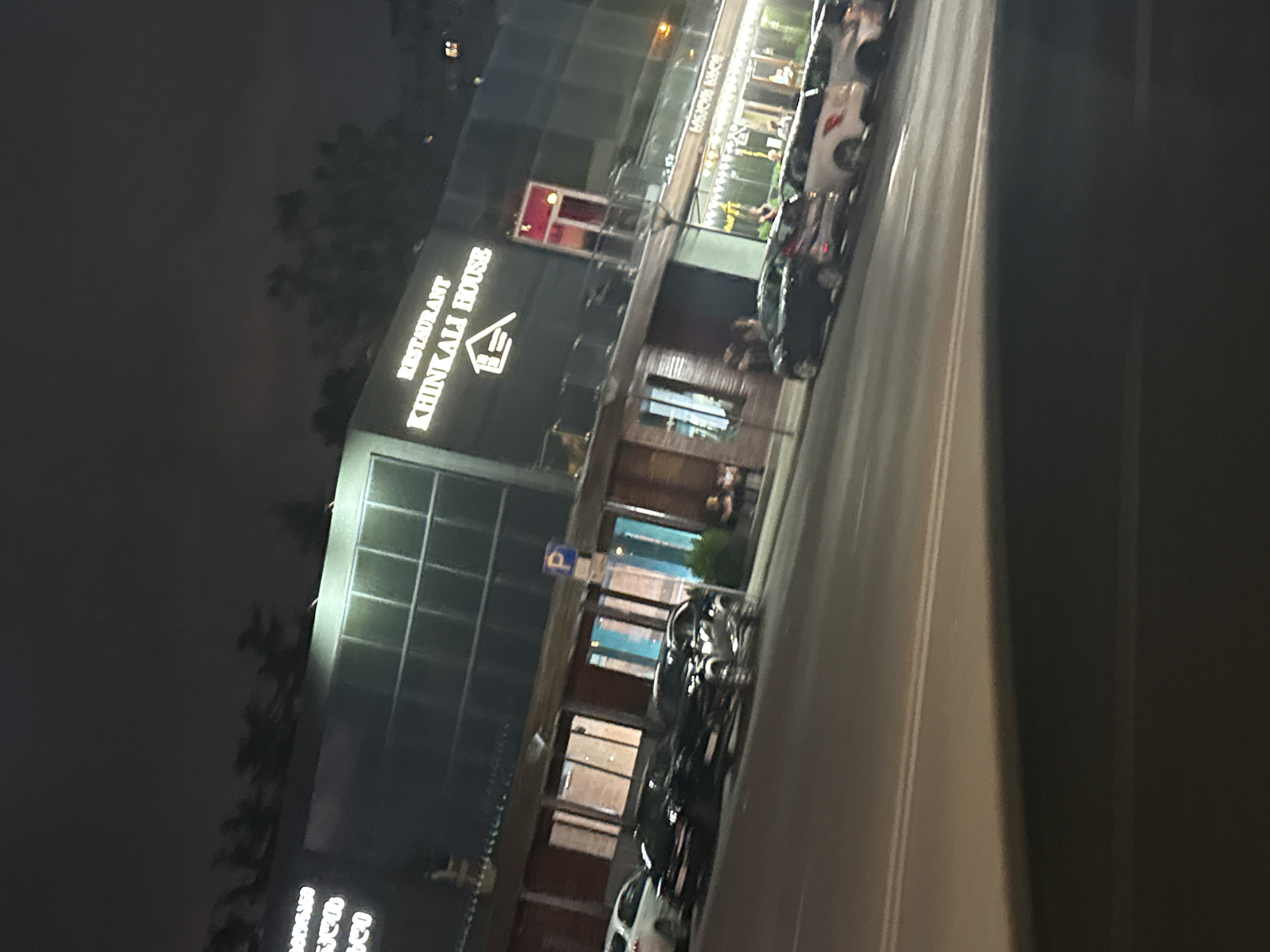 The image shows a nighttime street scene in Tbilisi, Georgia. On the right side of the frame is a building with a large illuminated sign that reads "RESTAURANT KHINKALI HOUSE". Below this sign, there is another illuminated sign in Georgian script. Several cars are parked along the street in front of the building, including a black SUV, a white sedan, and other vehicles. A blue parking sign with a white 'P' is visible on a pole. The street is paved, and a pedestrian walkway is visible behind the parked cars. The overall environment appears to be urban.
