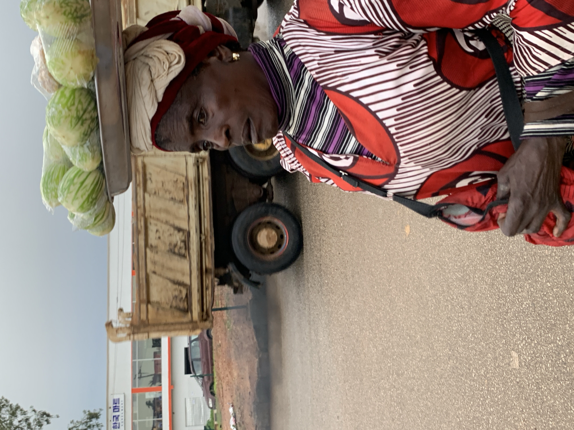 A medium-shot, eye-level, outdoor photograph captures a woman in profile, facing right, balancing a metal tray laden with green vegetables on her head. She wears a multi-colored striped top and a red and white patterned dress, with a red headscarf. A gold earring is visible in her right ear. In the background, a large yellow truck is parked, and a red car is visible to the left. A white building with Korean signage is behind the truck. The ground is a wet, paved surface, suggesting it may have rained recently. The scene appears to be in a semi-urban area, with elements of commerce and transportation. The location is identified as Gura Top, Nigeria.