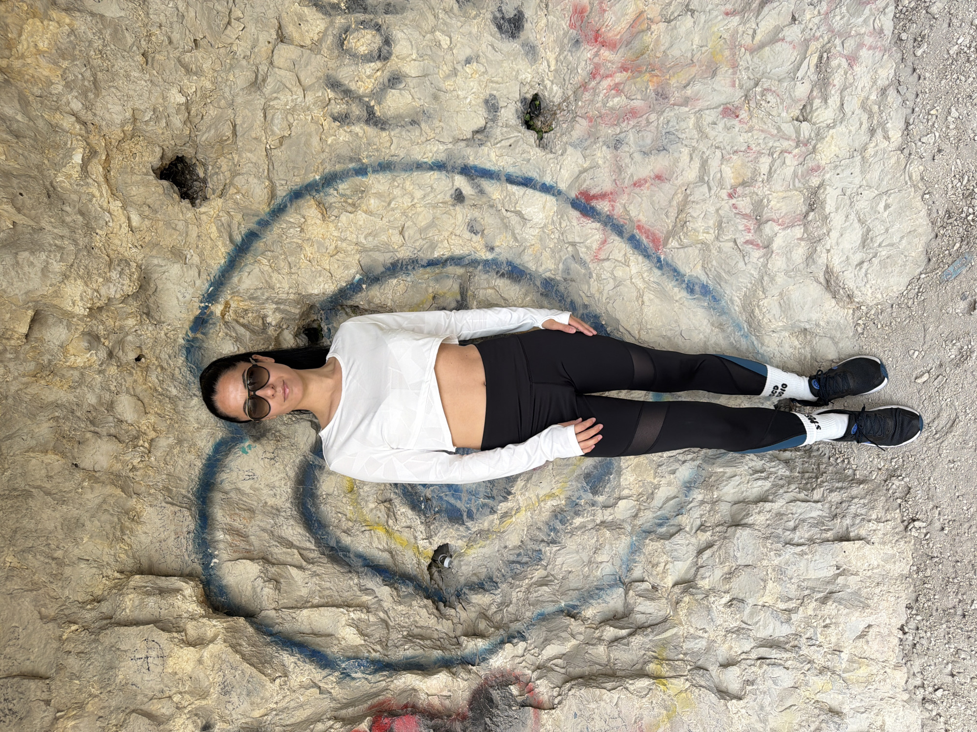 A woman is lying on a rocky, graffiti-covered surface. She is wearing sunglasses, a white long-sleeved crop top, black leggings with mesh panels, and black sneakers with white socks. The background is a textured, light-colored rock face with graffiti, including a blue circle. The image was taken in Pleven, Bulgaria.