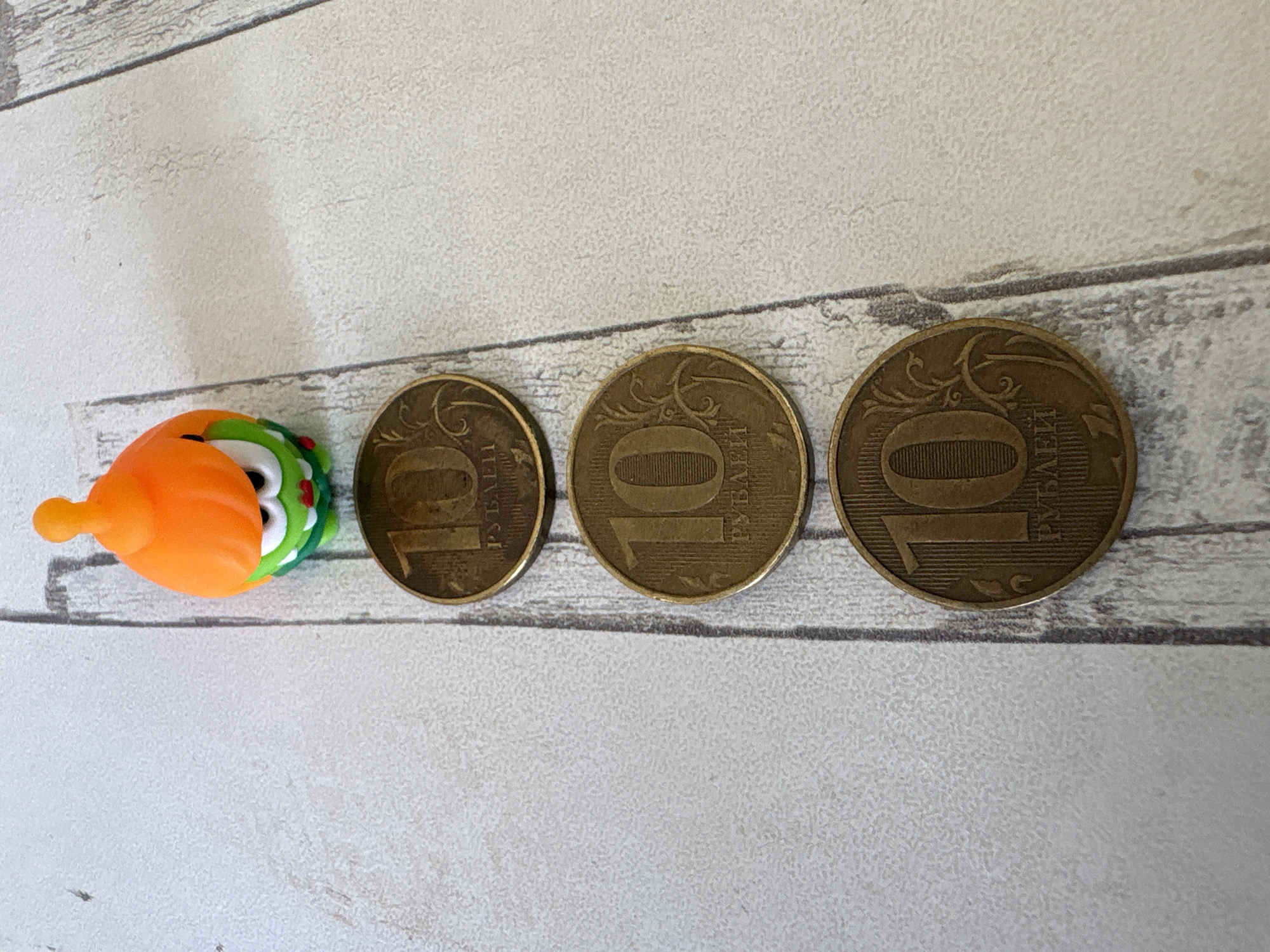 The image shows three 10 Russian Ruble coins, placed in a line from left to right. Next to the leftmost coin is a small, orange and green toy with white eyes. The items are situated on a textured surface that resembles white wood paneling. The context of the city of Chaikovsky, Russia is not discernible from the image.