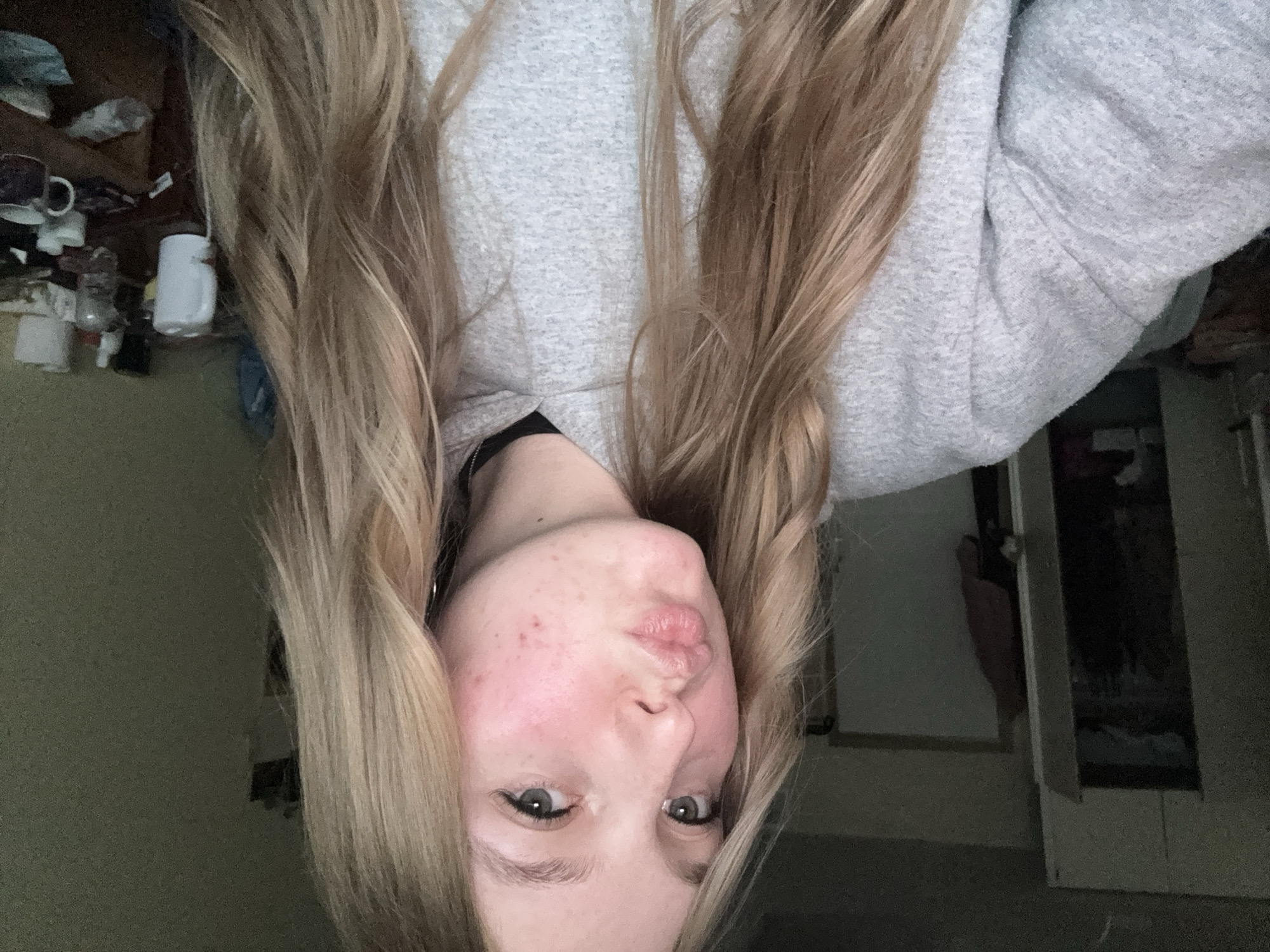 A casual selfie captures a young woman with light blonde or light brown hair, framing her face on both sides. She is looking directly at the camera with light-colored eyes, appearing to be blue or grey, and has eyeliner applied. Her lips are puckered in a playful "fish face" expression. Her cheeks appear slightly flushed or reddish, and some minor skin blemishes are visible on her chin and cheek. She is wearing a light grey hooded sweatshirt, with a glimpse of a black garment visible beneath her neckline.
The background suggests an indoor setting, likely a room within a home. To the woman's right (viewer's left), there is a cluttered surface or shelf holding various items, including multiple white and patterned mugs or cups, and bottles. To her left (viewer's right), a darker wall or corner is visible, along with a partially open wardrobe or cupboard revealing some contents. The room's walls appear to be a muted olive green or greyish-green color. The lighting is dim, indicating an indoor environment without strong natural light. The overall mood is informal and candid. No visible text is present in the image. The photo was taken in Perm, Russia.