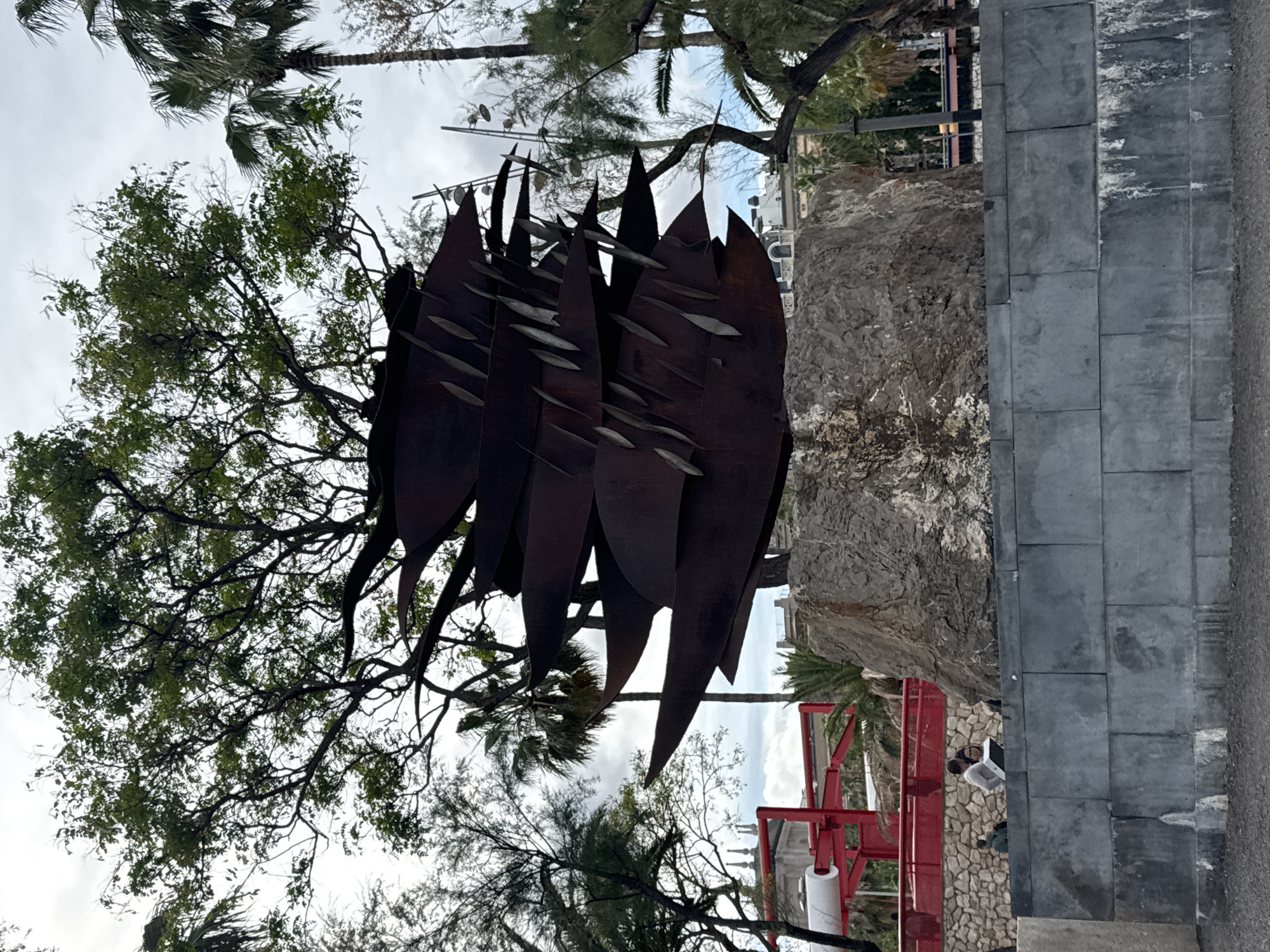 The image shows a public art installation in Barcelona, Spain. The sculpture is composed of large, dark metal "leaves" arranged vertically and clustered together. The leaves have a textured surface, appearing aged or rusted. In the background are trees with green foliage and palm trees, suggesting an outdoor environment. A large rock formation is visible behind the sculpture, along with part of a red mechanical structure. To the right of the rock is a gray stone wall. At the base of the wall is a person working with a laptop. The sky is overcast with a light cloud cover.