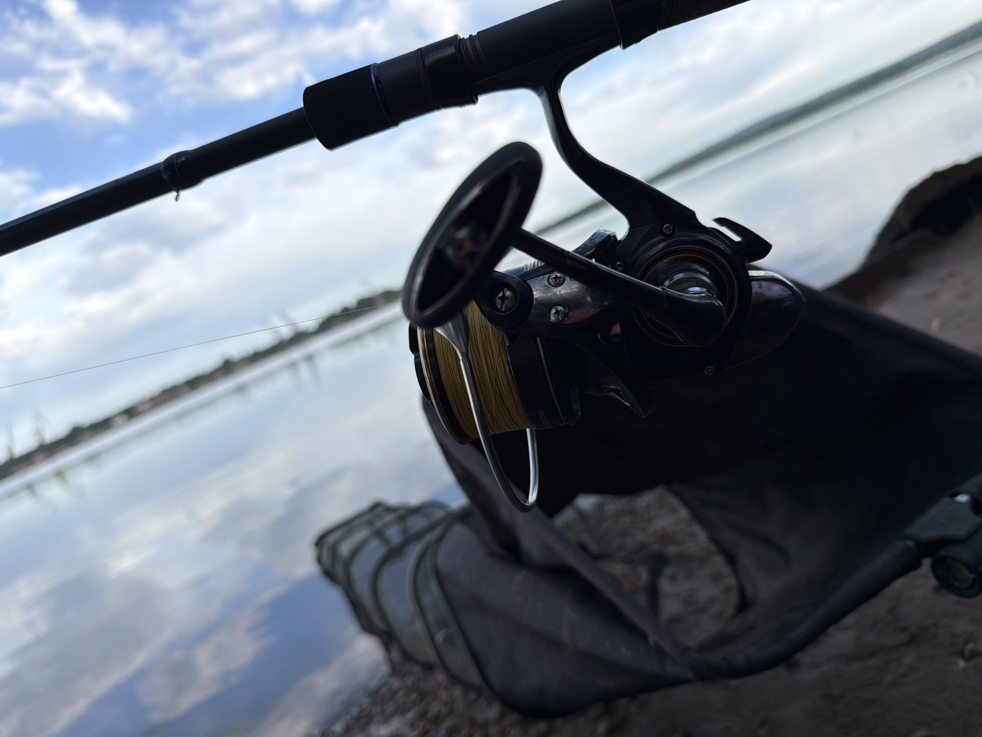 A close-up photograph shows a fishing rod and reel, resting on a dark-colored landing net on a gravelly surface near the water. The fishing rod is black with a blue accent, and the reel is silver and black with gold fishing line. A body of water reflects the sky, which is partly cloudy, in the background. Based on the information provided, the location is inferred to be Chaikovsky, Russia.