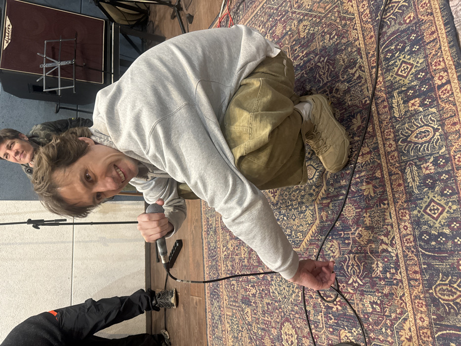 This image captures an indoor scene, identified as being in the city of Moscow, Russia, within what appears to be a recording studio or rehearsal space.
The central figure is a light-skinned male with brown, medium-length hair, positioned in a squatting stance on an intricately patterned rug. He is wearing a light grey hooded sweatshirt, olive-green loose-fitting pants, and tan athletic shoes over white socks. He holds a grey wired microphone in his right hand and smiles directly at the camera. His left hand is extended downwards, appearing to interact with black audio cables scattered across the rug.
In the mid-background, a light-skinned female with short, dark hair is partially visible behind the primary subject, looking slightly upward and to the right with a faint smile. She is dressed in a dark jacket. To the far left, the lower leg and foot of another individual are visible, wearing black cargo-style pants with an orange stripe and dark athletic shoes.
The environment features a dark wooden floor partially covered by a large, multi-colored Persian-style rug. The background wall on the left is lined with light-colored rectangular acoustic panels. Several black microphone stands are present, with at least two positioned against the acoustic panels. A dark-red amplifier or speaker cabinet, bearing the name "D'Addario," is visible in the background to the right, with a black music stand placed in front of it. Multiple black cables are visible on the rug and floor, indicative of audio or power connections.