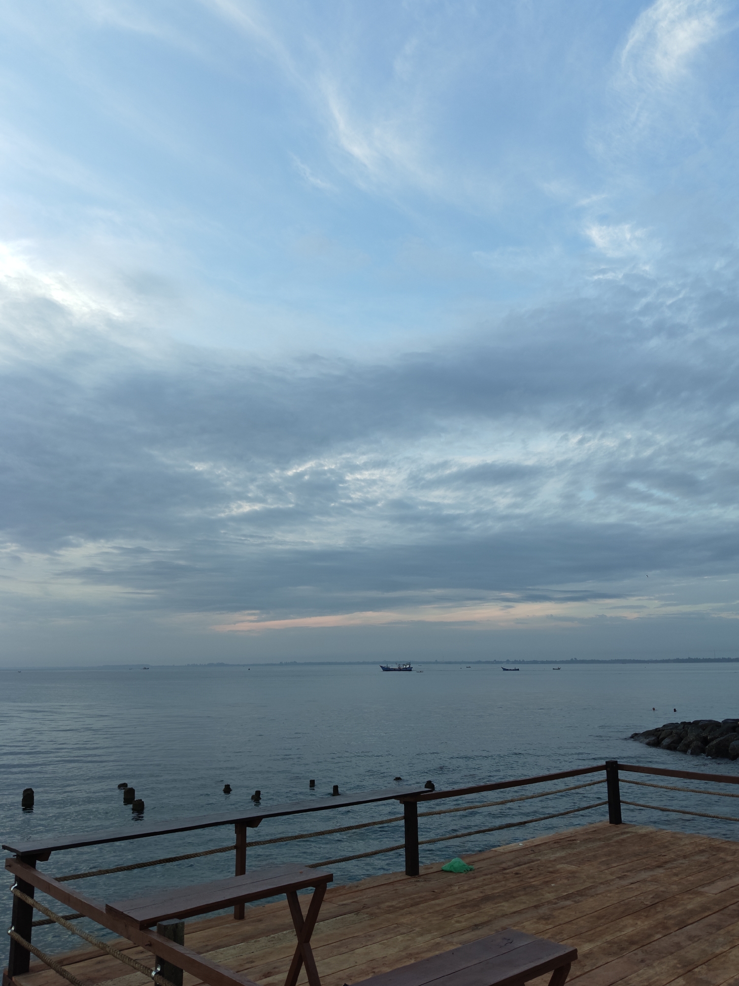 A wide-angle, eye-level photograph captures an early morning or late afternoon scene at a wooden pier overlooking the ocean in Lhokseumawe, Indonesia. The sky is a soft gradient of pale blue and grey, with wispy clouds scattered across the expanse. A faint blush of pink is visible near the horizon line, suggesting the soft light of sunrise or sunset. The ocean is calm, with gentle ripples on its surface. Several small boats are dotted across the water in the distance.
The foreground of the image features a wooden deck with a rustic wooden bench and table set. A rope railing, held up by dark wooden posts, separates the deck from the water. In the water just beyond the pier, several weathered wooden pilings emerge from the surface. To the right of the deck, a rocky breakwater extends into the sea. The overall atmosphere is peaceful and serene.