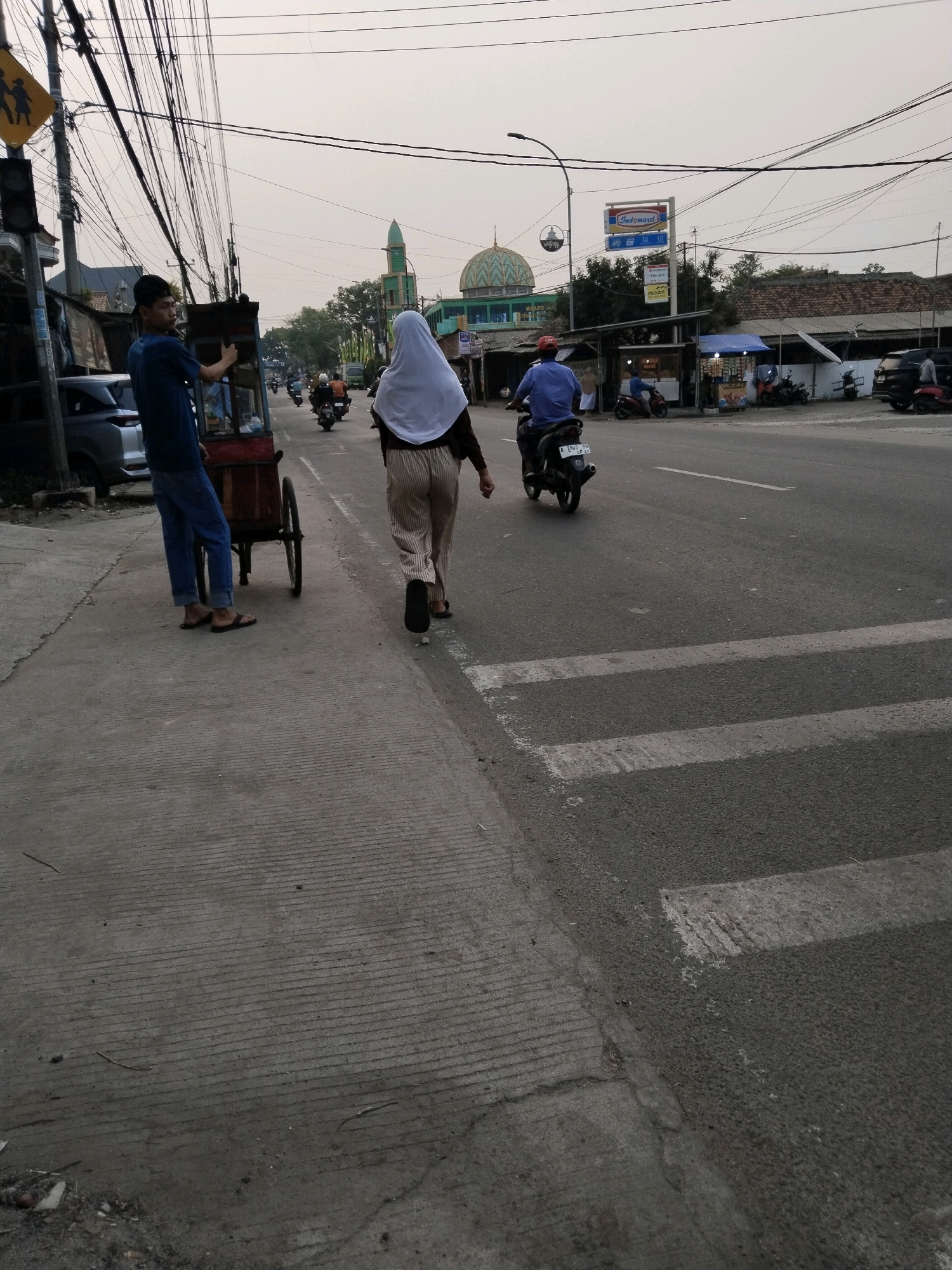 In the city of Serang, Indonesia, a pedestrian crosses a street with a marked crosswalk. To the left, a man wearing a blue shirt and jeans stands on the sidewalk next to a food cart. Further down the sidewalk, a woman in a white hijab and striped pants walks away from the camera, also heading towards the crosswalk. The street is lined with buildings, including a mosque with a green dome, and several shops. Vehicles, including motorcycles and cars, are visible on the road. The sky is overcast, and the overall lighting suggests it is either early morning or late afternoon. The scene depicts a typical urban streetscape with everyday activities taking place.