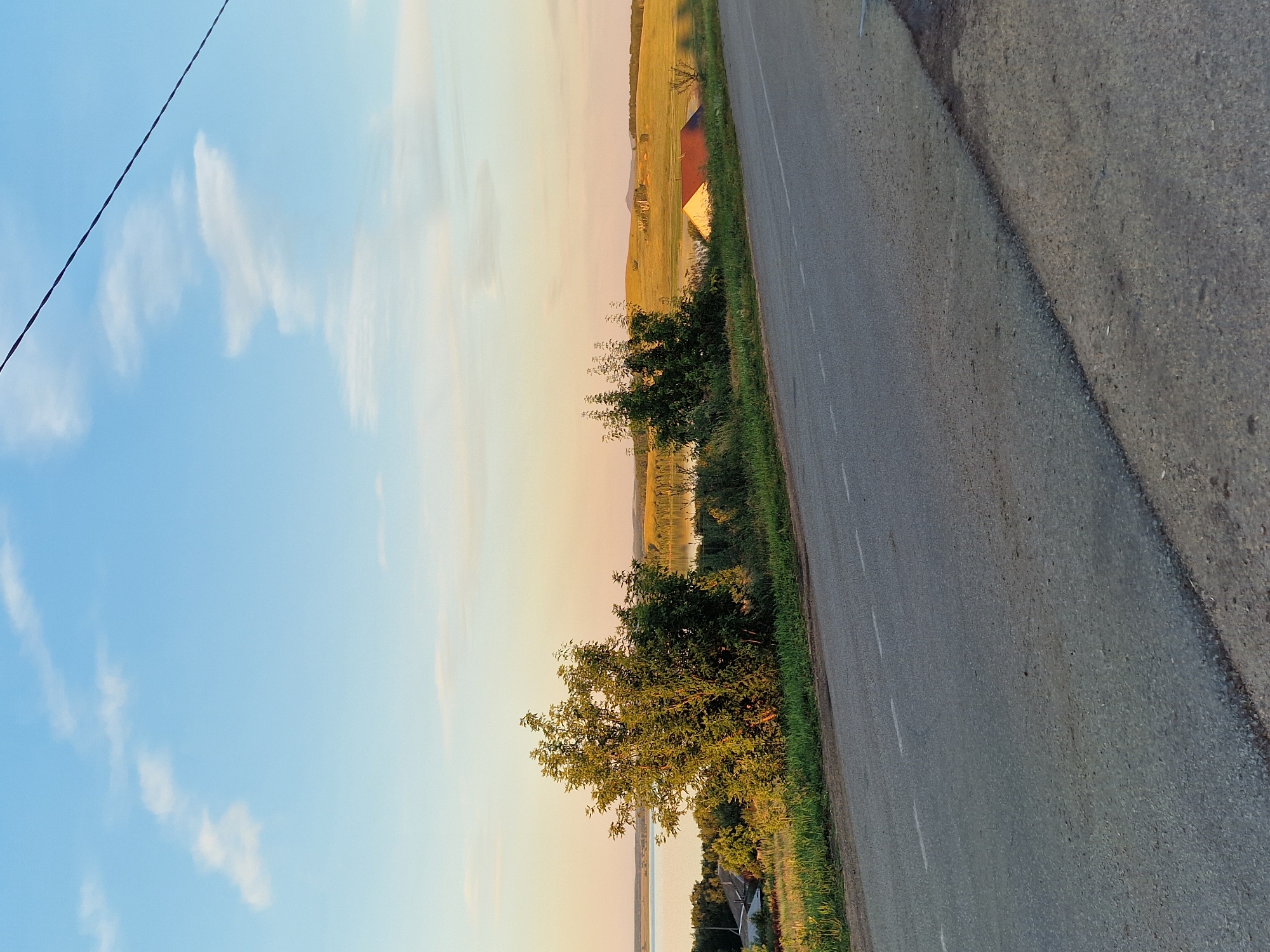 This is a photograph of a rural landscape with a road in the foreground. The road is paved and has dashed white lines marking the lanes. To the left of the road, there is a grassy embankment with some trees and vegetation. In the middle ground, there is a body of water, likely a lake or river, with a small village or settlement on its shore. Beyond the water, rolling hills are visible, with some scattered trees and buildings. The sky is clear with a few wispy clouds, and the overall lighting suggests it is either early morning or late afternoon. The image does not depict any people or specific actions. The location is not the city of Kashgar City, China.