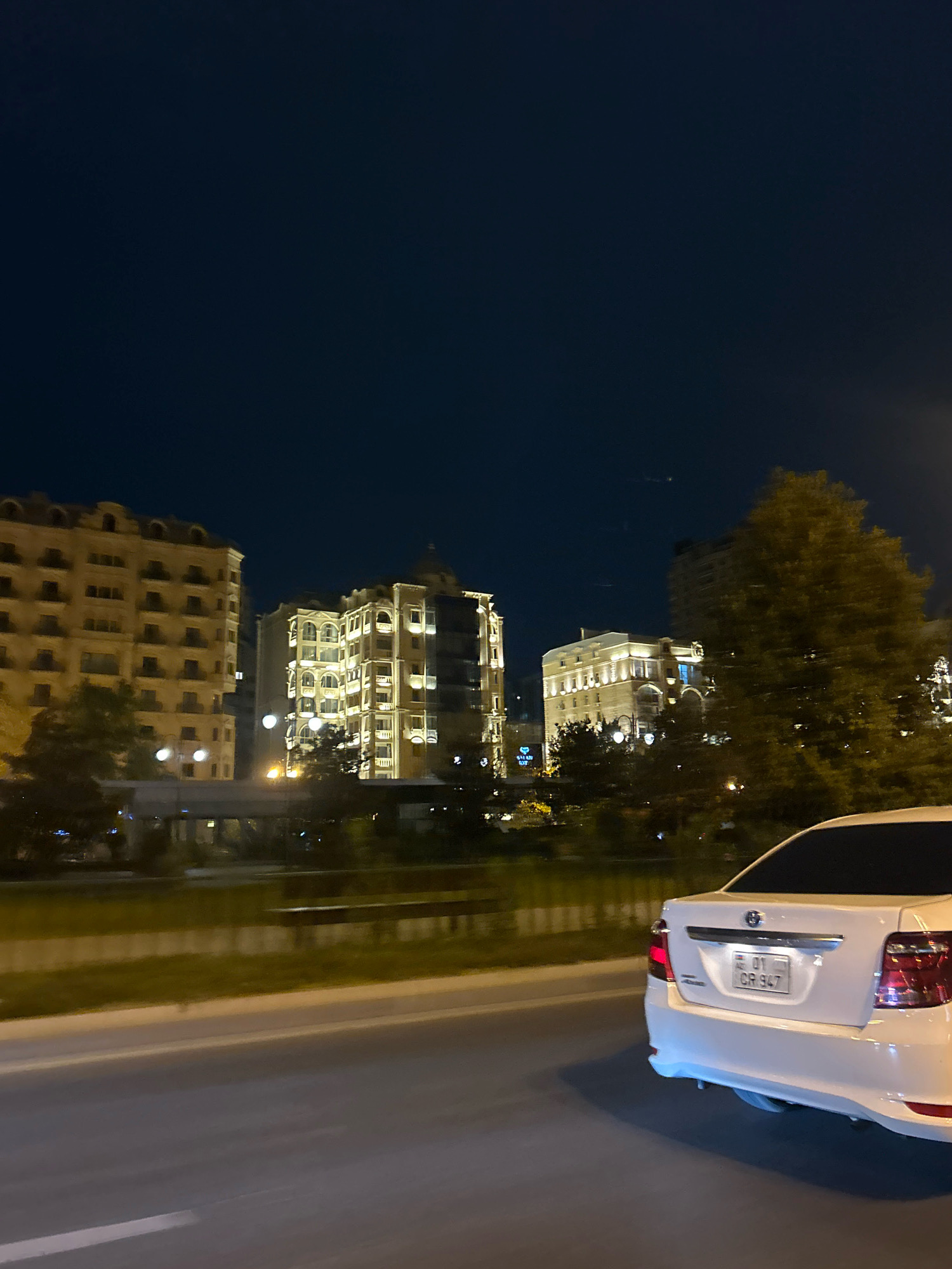 The image depicts a night-time view of the city of Baku, Azerbaijan, likely from a moving vehicle. The foreground shows the rear of a white sedan, identifiable by its license plate which reads "01 CR 947". The car is on a paved road, suggesting it is a street or highway.
In the mid-ground, a grassy median separates the road from the buildings. The buildings themselves are illuminated and appear to be apartment complexes or office buildings with distinctive architectural features, including numerous windows and decorative elements. Trees are also visible, some of which are illuminated by the building lights.
The background is dominated by a dark, clear night sky. The overall impression is of an urban environment at night, with modern architecture and street lighting. The specific location is difficult to pinpoint precisely without more context, but the architecture and the license plate format are consistent with Baku, Azerbaijan. There are no people visible in the image.