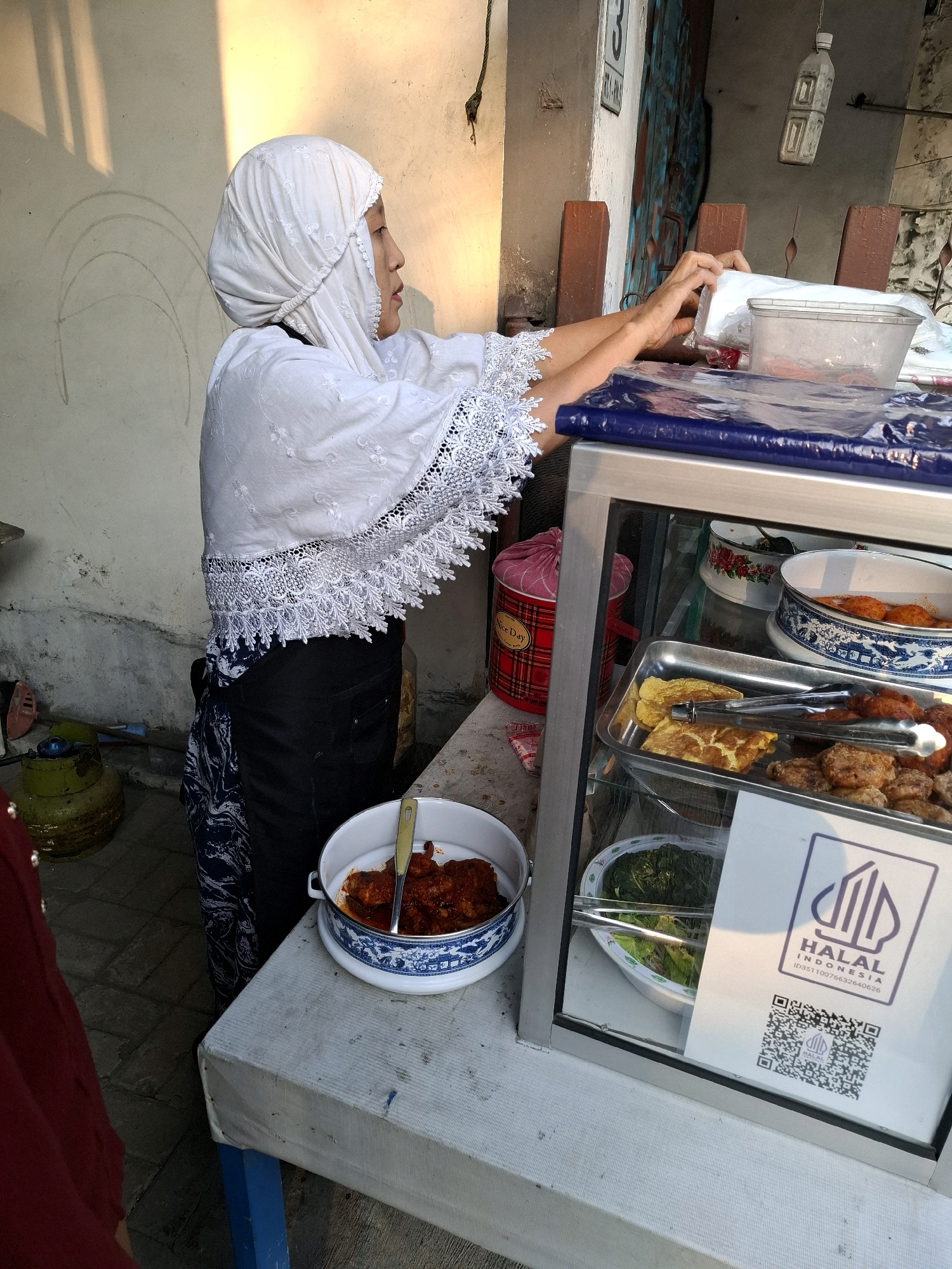 The image depicts a street food stall in Surabaya, Indonesia. A woman, dressed in a white headscarf and white embroidered blouse over a black skirt and patterned fabric, is seen from the side, reaching for a plastic container on a blue tarp-covered surface. In front of her, a glass display case reveals various food items. A metal tray contains what appears to be fried chicken, omelets, and other fried dishes, with tongs resting on the food. Below this, a dish of green vegetables is visible. A prominent sign with a "HALAL INDONESIA" logo and a QR code is displayed inside the case. To the left of the stall, a white table is partially visible, holding a blue and white patterned bowl filled with red-spiced food and a yellow serving spoon. A green gas cylinder is situated behind the table. The background shows a textured wall and wooden posts, suggesting an outdoor urban setting. The lighting indicates it is daytime, with sunlight casting shadows on the wall.