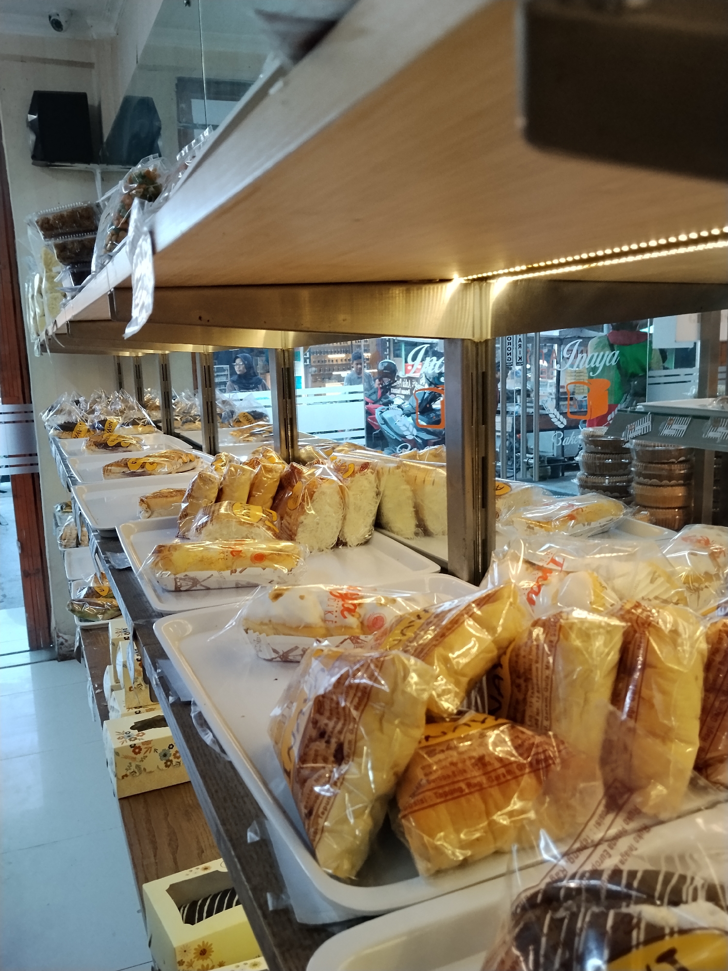 This is a medium-shot, eye-level, indoor photograph of a bakery display in Ciruas, Indonesia. The foreground features several rows of baked goods, primarily bread and cakes, packaged in clear plastic. The items are arranged on white plastic trays, which are placed on dark brown shelves. The lighting appears to be artificial, with a strip of small lights illuminating the underside of the upper shelf. The background shows the interior of the bakery, with reflections in a glass partition. Through the glass, we can see more baked goods, stackable containers, and a glimpse of a street scene with people and a motorcycle. There are also some decorative boxes and a CCTV camera visible on the upper left wall. The overall impression is of a well-stocked, commercial bakery.