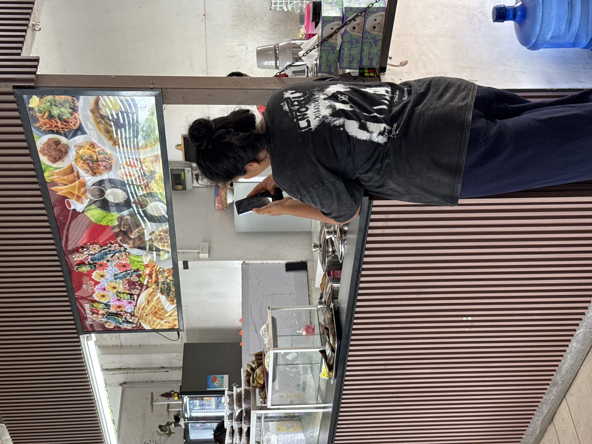 The media file depicts a rear view of a young woman with her hair in a bun, standing in front of a food stall in Baan Bo Phut, Thailand. She is wearing a dark, graphic t-shirt and dark pants, and is looking down at her smartphone. The stall displays a variety of food items on a large illuminated menu board. Visible food items include noodles, rice dishes, and what appear to be fried snacks. The setting is an outdoor or semi-outdoor market, suggested by the striped awning and the overall casual atmosphere. A large blue water dispenser is visible in the background. The time of day appears to be daytime. The image is oriented vertically.