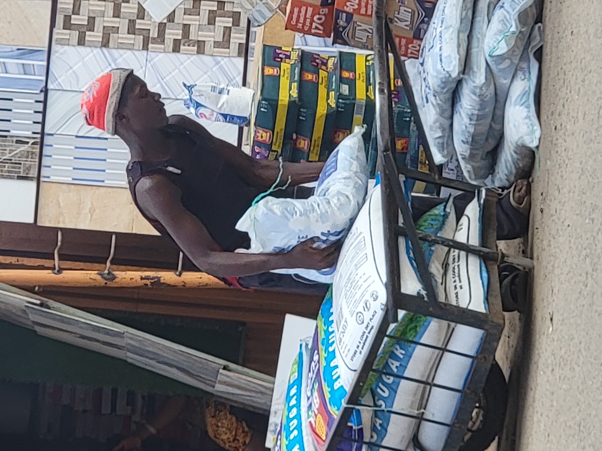 The image depicts a vibrant street vending scene in Maiburiji, Nigeria, under bright daylight conditions. A young man, with dark skin, wearing a dark sleeveless top and a distinctive orange and white knitted cap, stands beside a metal pushcart. He is looking upwards and appears to be holding a white bag, seemingly engaged in selling or arranging merchandise.
The pushcart is heavily loaded with various goods. Prominently displayed are multiple large white and blue bags, identifiable as sugar, with text such as "SUGAR" and "BUA SUGAR," along with storage instructions "STORE IN A COOL DRY PLACE." Above these, several yellow and green boxes are stacked, bearing labels like "34 sachets," "170g," and the brand name "Klin."
In the background, a wall is visible, decorated with an assortment of ceramic tiles featuring geometric, striped, and plain patterns, suggesting the exterior of a building or a robust market stall. Below the tiled wall, a wooden beam structure can be seen. The strong, natural sunlight indicates a clear and sunny day, enhancing the details of this bustling outdoor market environment.