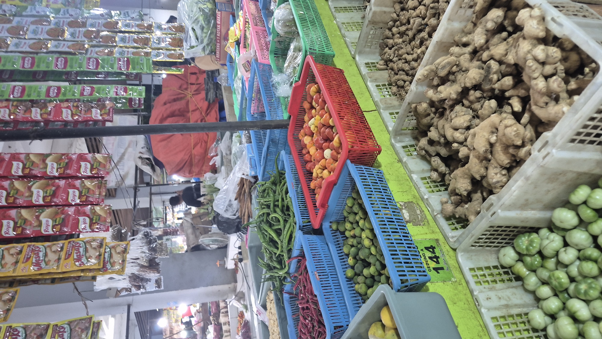 This is a close-up, eye-level, medium shot of a market stall in Bojonegoro, Indonesia, displaying an assortment of fresh produce and packaged goods. In the foreground, several white plastic crates are filled with various items. The left-most crate contains small, pale green tomatoes. To its right, another crate holds a large quantity of ginger roots, with their earthy brown skin and knobby texture. Further to the right, partially visible, is another crate filled with ginger. In the midground, there are several colorful plastic baskets. A bright red basket is filled with small, round red and yellow tomatoes. Adjacent to it, a blue basket contains a dense pile of small, green, round fruits, possibly limes or small tomatoes. Behind these, a blue basket is overflowing with long, dark green chili peppers. To the left of the chili peppers, another blue basket holds thin, dark red chili peppers. In the background, hanging from overhead structures, are numerous packets of seasonings and spices, predominantly in red and yellow packaging, some displaying the brand name "Royco." A tall, dark pole, likely part of the stall's structure, bisects the frame vertically. Out of focus in the far background, a person wearing a dark shirt is visible, and beyond them, various market activities are suggested, including other stalls and a large red tarp. The overall environment appears to be an indoor market with overhead lighting, casting some shadows. A green sign with white lettering, partially visible in the lower left quadrant, reads "GEDUNG A 144."