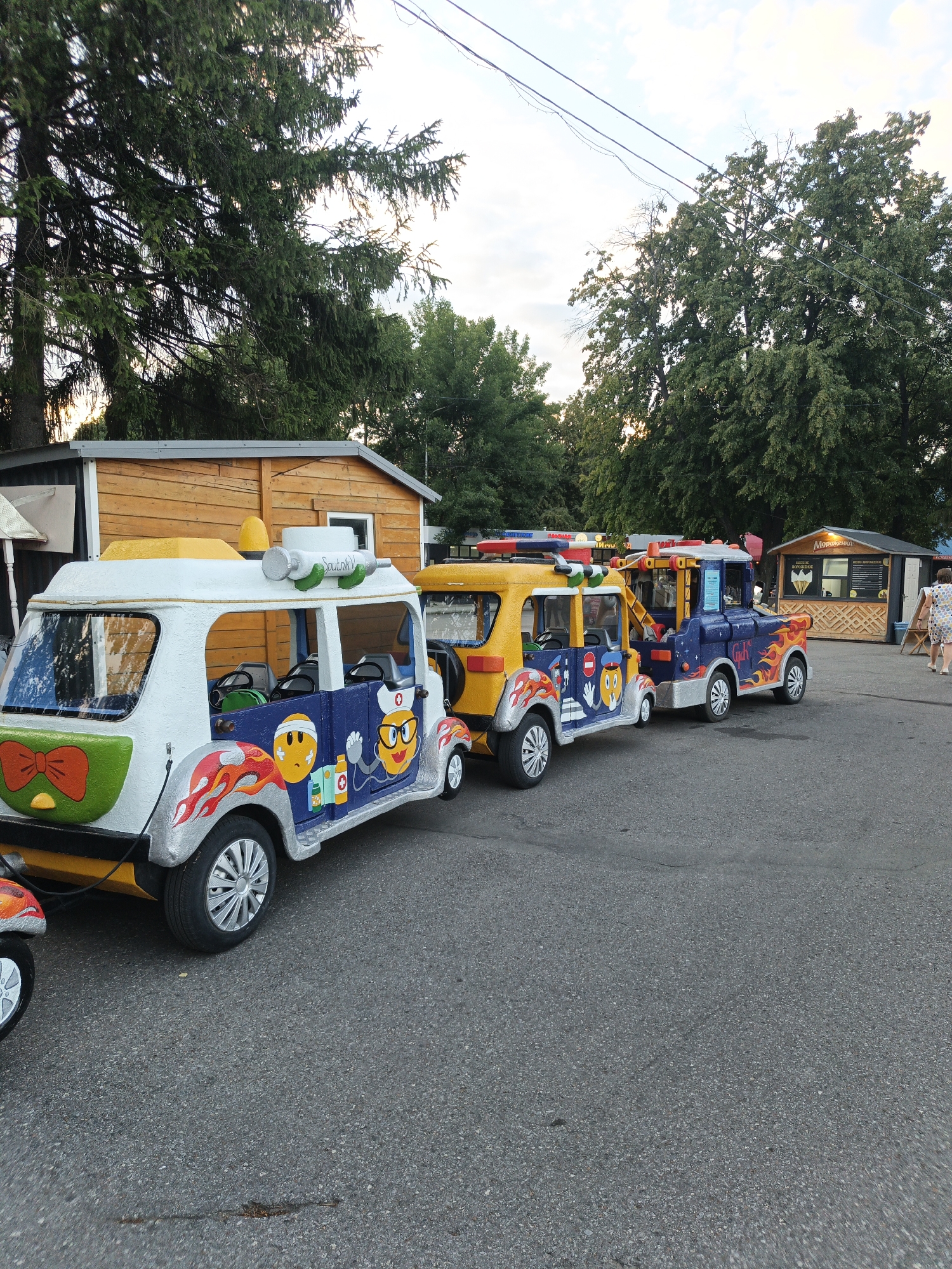 The image shows a line of colorful, cartoon-themed kiddie rides shaped like vehicles, parked on asphalt. The first ride, in the foreground, is white and blue with a large orange bow on the back and flames painted on its sides. It features cartoon characters with smiley faces, one dressed as a doctor. On its roof, there is a yellow flashing light and a large syringe labeled "Sputnik." To its right is a yellow vehicle with similar flame designs and cartoon characters, followed by a blue vehicle resembling a tow truck, also adorned with flames. These rides appear to be in an outdoor amusement area or park, with trees and a wooden building in the background. The wooden building has a sign with Cyrillic text that is partially visible. Further in the background, another small wooden structure with a sign displaying "Мороженое" (Ice Cream in Russian) and an ice cream cone graphic suggests a food vendor. A woman in a light-colored dress is walking away from the camera in the background. The lighting suggests it is daytime, possibly late afternoon or early evening, with a slightly overcast sky. The overall atmosphere is playful and geared towards children's entertainment.