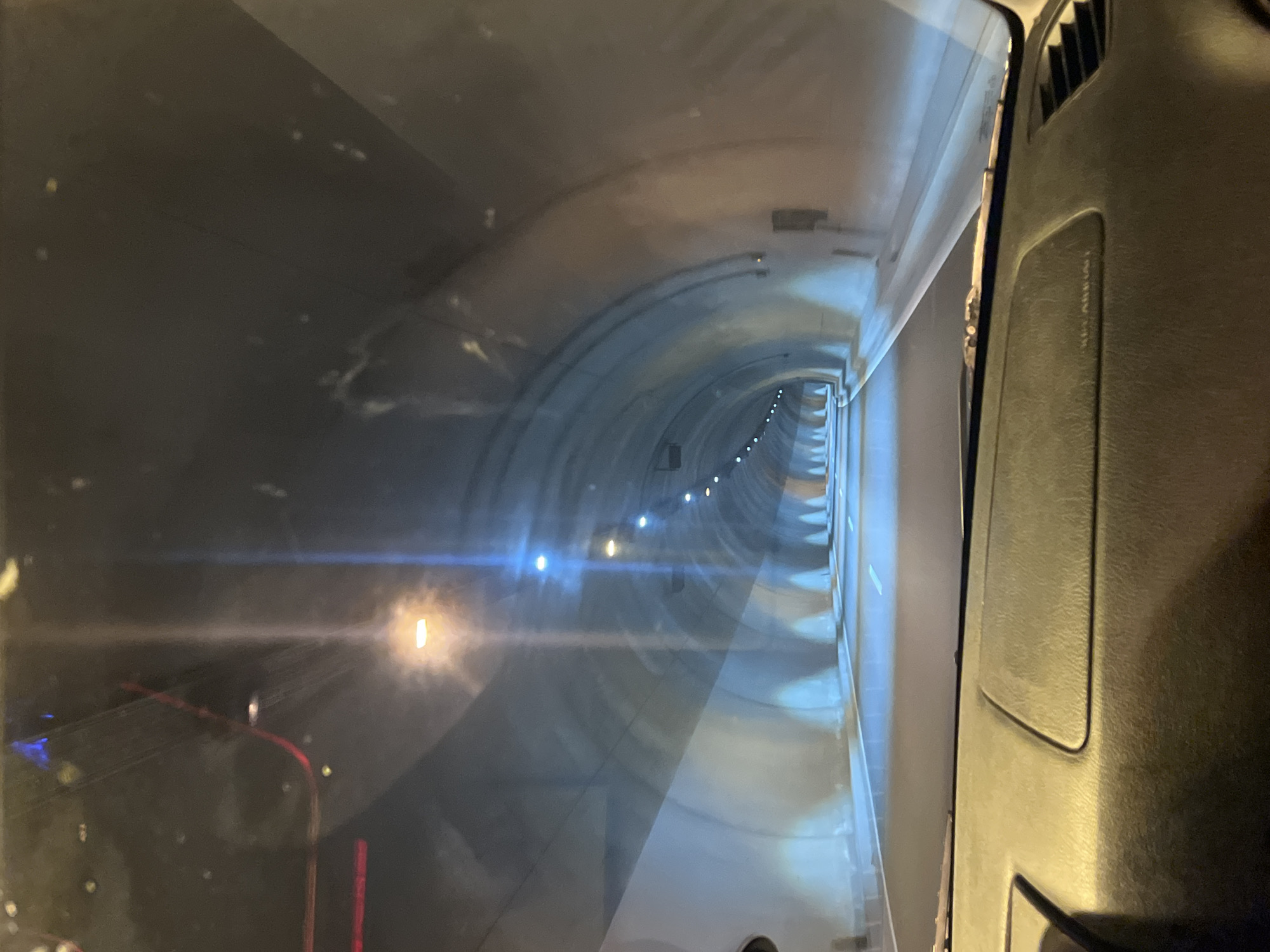 The image shows the interior of a vehicle driving through a tunnel. The perspective is from the driver's seat, looking forward through the windshield. The tunnel appears to be well-lit with overhead lights and lights along the tunnel walls. The road surface is visible, with white lane markings. The interior of the car, including the dashboard, is also visible in the foreground. The scene suggests movement through an enclosed passage, typical of a road tunnel. There are no people or specific landmarks visible that would indicate a location in Blatino, Bulgaria.