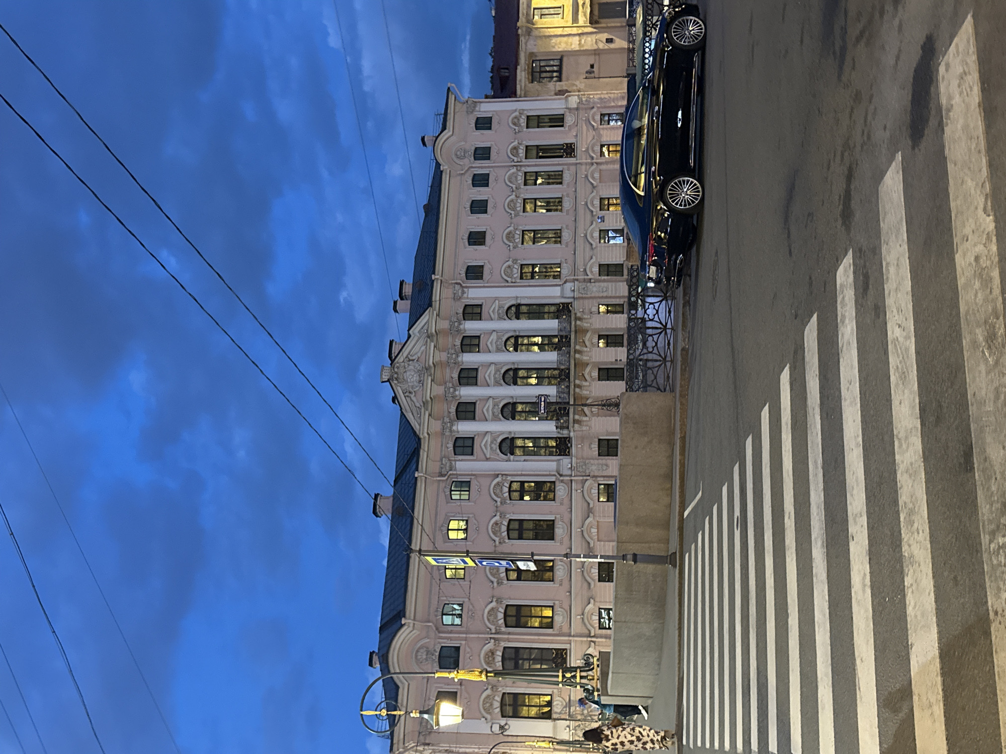 The image, correctly oriented, captures a twilight or nighttime street scene in Saint Petersburg, Russia. A grand, multi-story building with a light pink facade stands prominently on the left. It features intricate architectural details, including white decorative columns, cornices, arched windows, and small balconies. Many of the building's windows are illuminated, casting a warm glow.
Above the building, the sky is a deep blue, scattered with darker clouds, and several thin utility wires stretch across the frame. On the right, a dark car is parked along the curb, alongside a metal fence. In the foreground, a prominent zebra crossing with broad white stripes spans the dark asphalt street. Shadows, likely cast by the streetlamp, stretch across the pedestrian path. A decorative streetlamp with golden elements and a lit bulb is visible in the lower center, and part of a person's lower body, dressed in patterned clothing, is just visible at the bottom right corner near the streetlamp's base. A small blue street sign with white, illegible Cyrillic-like characters is attached to the building facade. The scene conveys a quiet, urban evening ambiance.