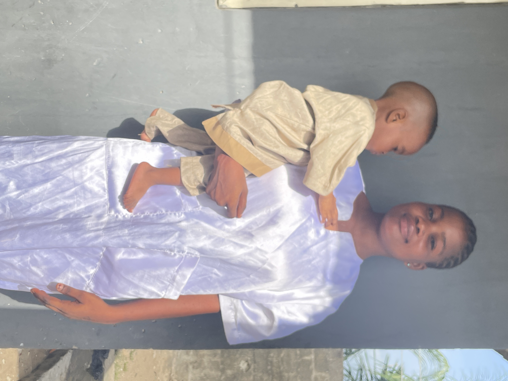 An outdoor image, likely taken in Badagry, Nigeria, shows a woman and a young child interacting playfully under bright sunlight. The woman, with dark skin and braided hair, is positioned upright, looking directly at the camera with a clear smile. She wears a long, flowing white garment made of a shiny, possibly satin, fabric. A small, light-colored earring is visible in her right ear, and her right hand, with a hint of red nail polish on the thumb, is visible near her waist.
A young child, appearing to be a toddler, is situated on the woman's front. The child wears a light tan or beige outfit. Their bare feet are placed on the woman's stomach or hip area, and their hands are holding onto her upper chest. The child's head is turned downwards and away from the camera, making their expression unclear.
The setting appears to be outdoors, with a plain, greyish surface forming the background and ground. Bright sunshine casts distinct shadows, indicating a clear, sunny day. A glimpse of green foliage, possibly palm leaves, can be seen in the bottom right corner of the image. The scene captures a moment of affectionate playfulness between the woman and the child. No visible text is present in the image.