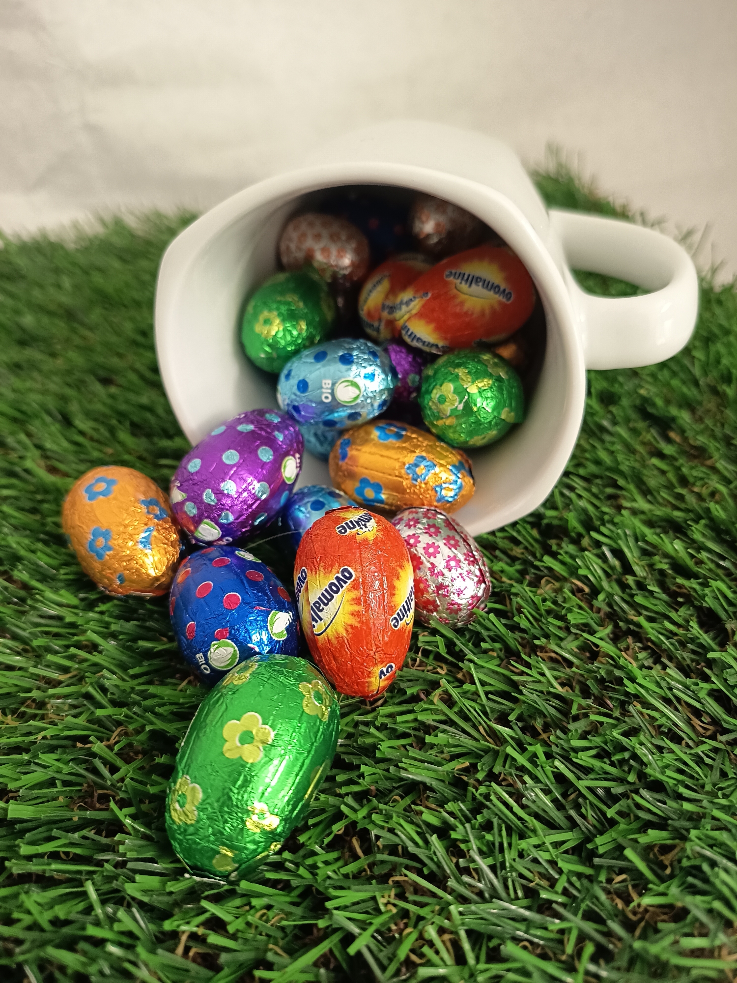 A tipped-over white mug spills out colorful foil-wrapped chocolate Easter eggs onto artificial green grass. The eggs display various colors and patterns, including floral designs, polka dots, and brand names like "Ovomaltine" and "Bio." The background features a blurry white surface. The photo does not display any humans, subjects, actions or interactions. It is impossible to ascertain whether the image was taken in Wangen bei Olten, Switzerland, without additional information.