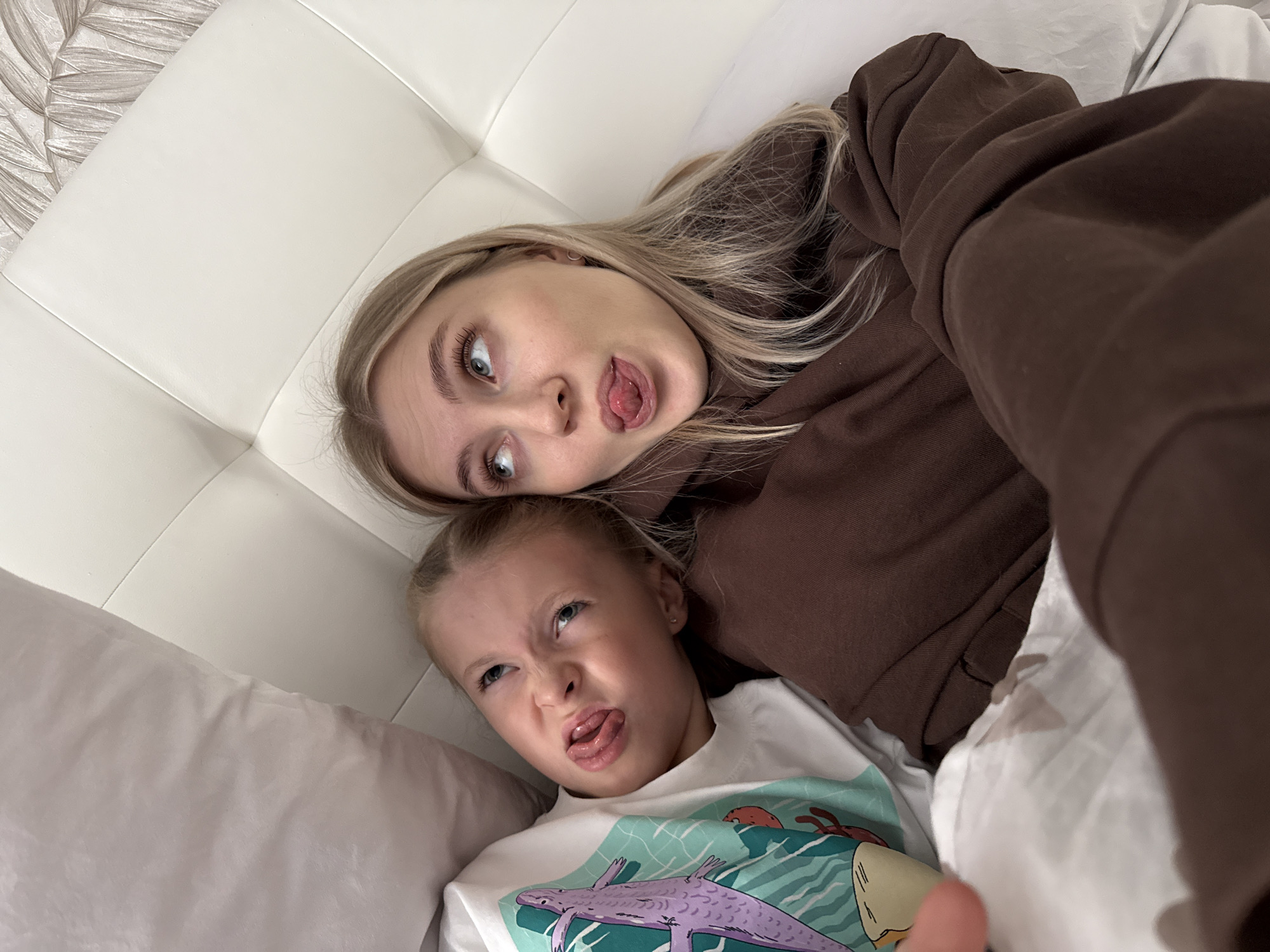 Two females are lying on a white bed. An older female, with blond hair, wears a brown sweatshirt and is above a younger female wearing a white t-shirt with an image of a purple lizard. Both females are sticking out their tongues and making silly faces. A white headboard is visible behind the older female. The location is a bed, not explicitly in Izhevsk, Russia.