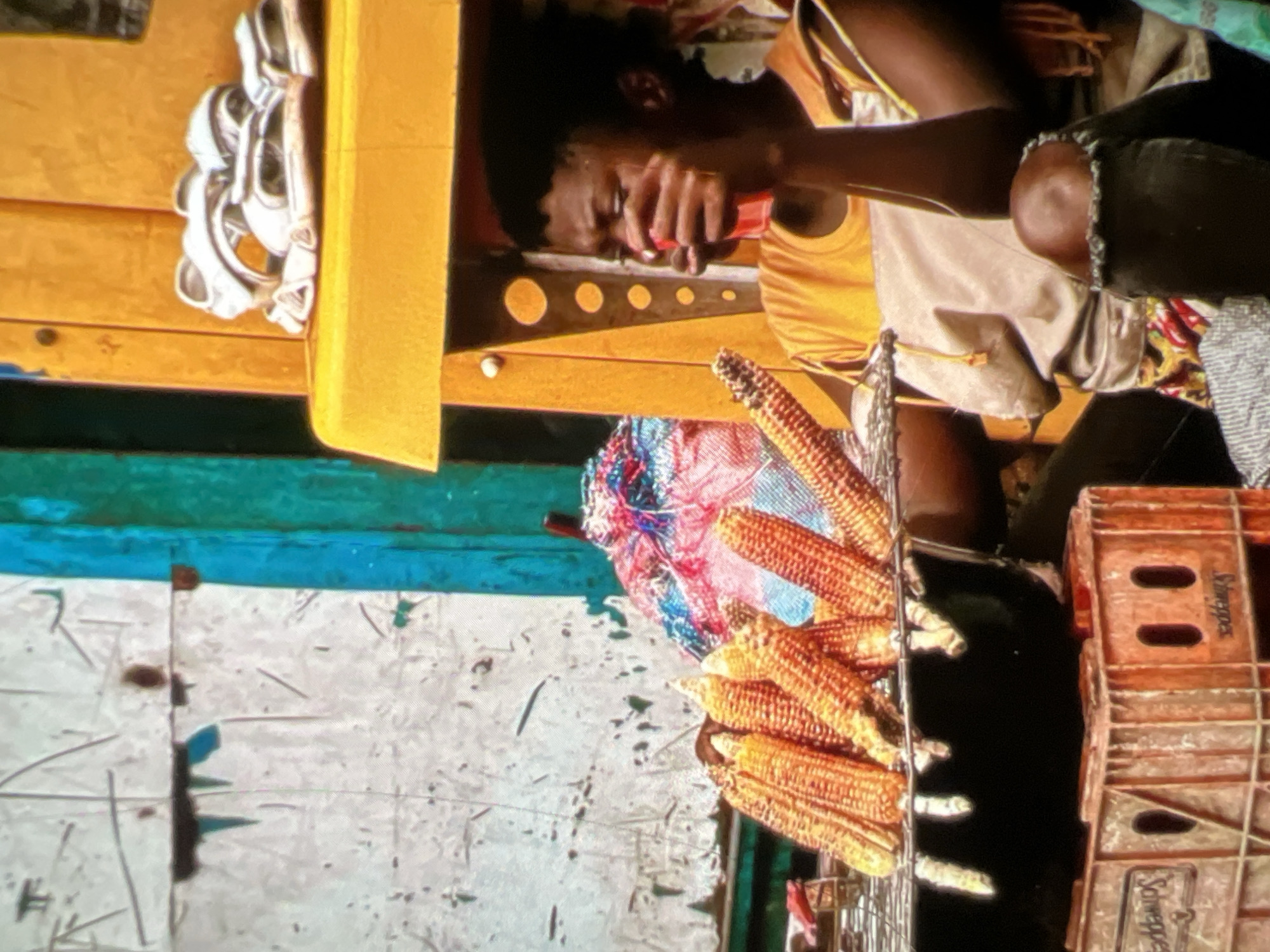 In Lagos, Nigeria, a young person is captured in a candid moment, seemingly taking a break from their vending duties. They are seated with their legs crossed, dressed in a casual orange t-shirt and dark, ripped jeans, with a light-colored fabric draped over their lap. Their hand is raised to their mouth, holding a red object, possibly a drink or a snack. The surrounding environment is a vibrant street scene. In the foreground, a wire rack holds several ears of corn, suggesting they are selling the produce. Beneath the rack sits a black pot, and below that, orange plastic crates branded with "Schweppes" are stacked. To the right, a bright yellow structure with circular cutouts serves as a backdrop, where a pair of white sandals rests. The wall behind them is a textured, weathered surface with green and blue painted elements. A colorful, patterned fabric bundle is visible to the left of the yellow structure.