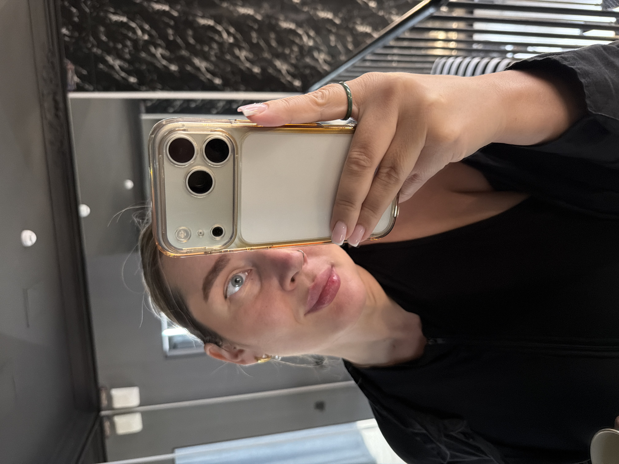 A person is taking a selfie using a gold-cased iPhone. The individual has light skin, blue eyes, and light-colored hair. They are wearing a black top. A green ring is visible on their finger. The background appears to be a bathroom, with a marble wall and a mirrored surface. The location is unknown.