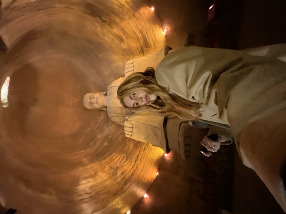 The image depicts a female subject posing within the Shizhong Mountain Grottoes in Shizhong, China. She is situated in the foreground, slightly right of center, gazing directly at the camera with a subtle expression. She has long, wavy, light-colored hair and is wearing a light beige, oversized coat. Her left hand, adorned with several rings, holds a dark, rectangular object. Behind her, a large, light-colored stone Buddha statue is prominently seated in a meditative pose. The environment is a dimly lit, cavernous space with textured, reddish-brown rock walls that arch overhead, indicative of a carved grotto. Multiple small, warm-toned string lights are visible along the lower sections of the cave walls, casting a soft, orange-yellow glow throughout the scene.