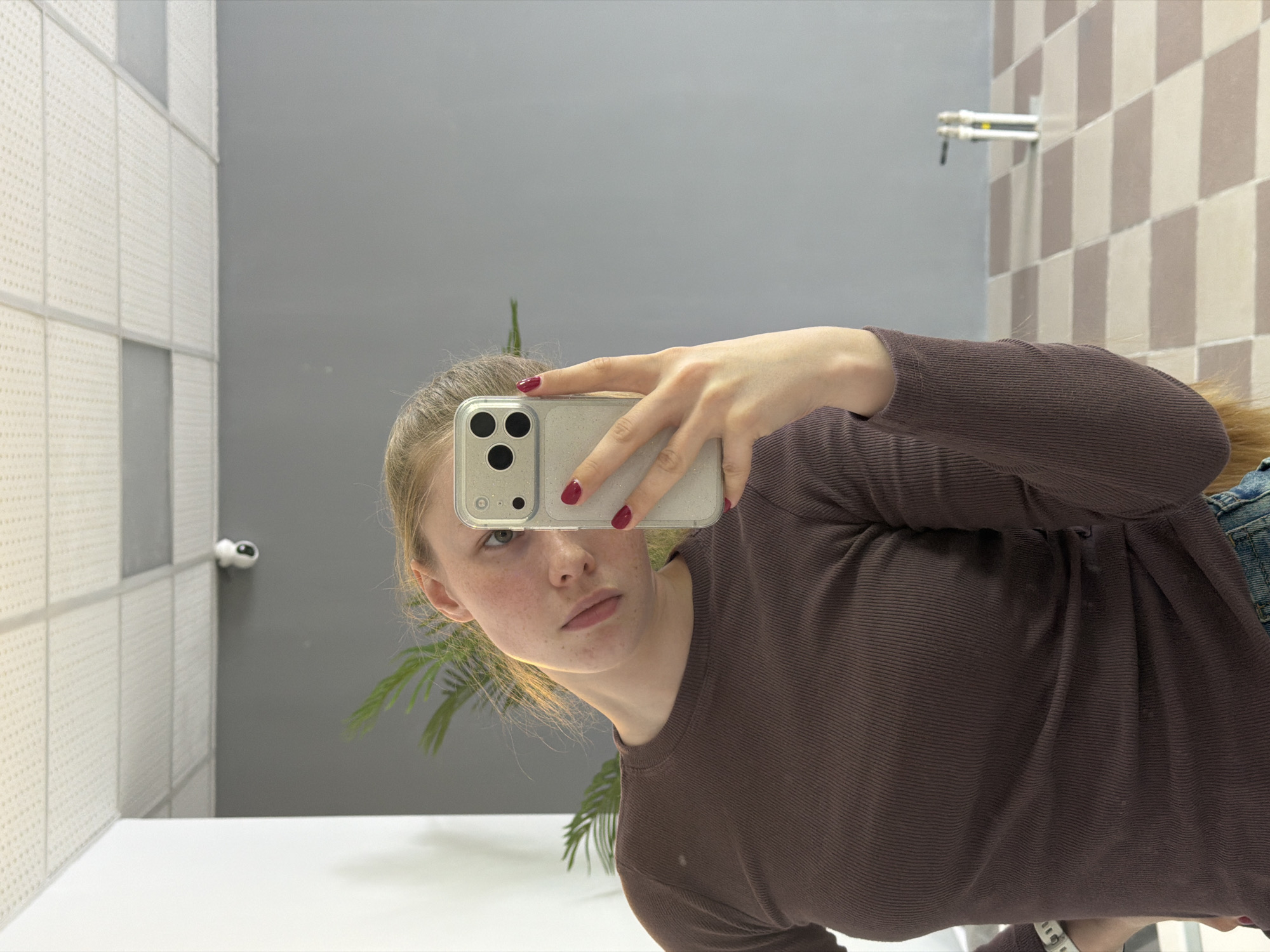 A selfie taken in a bathroom setting. The subject is a young woman with fair skin and light hair, wearing a brown ribbed long-sleeve shirt. She is holding a smartphone up to take the picture, and her red nail polish is visible on her fingers. The background reveals a tiled wall, a grey wall, and what appears to be a white sink or countertop. There's also a plant reflection and some bathroom fixtures visible. The location is not specified or discernible from this image.