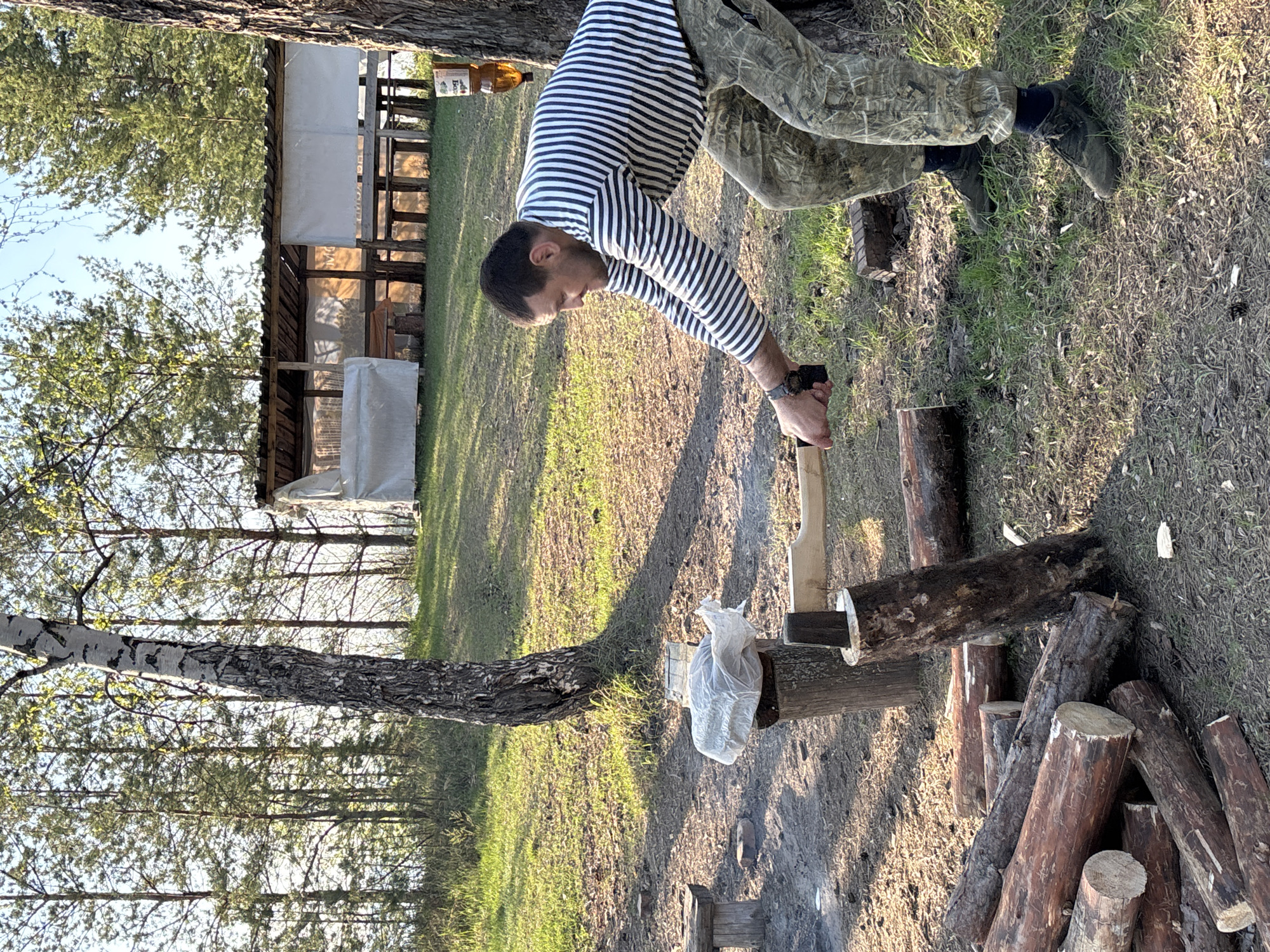 In Vekoshinka, Russia, a man in a striped shirt and camouflage pants is splitting wood outdoors. He is bent over a log with a hatchet in his hands. A pile of logs sits on the ground near him. A tree trunk lies horizontally across the frame, and a plastic bag sits on a log next to the tree trunk. A wooden structure is visible in the background, partially covered with white fabric. The ground is covered in grass and wood chips, and trees are in the background under a blue sky.