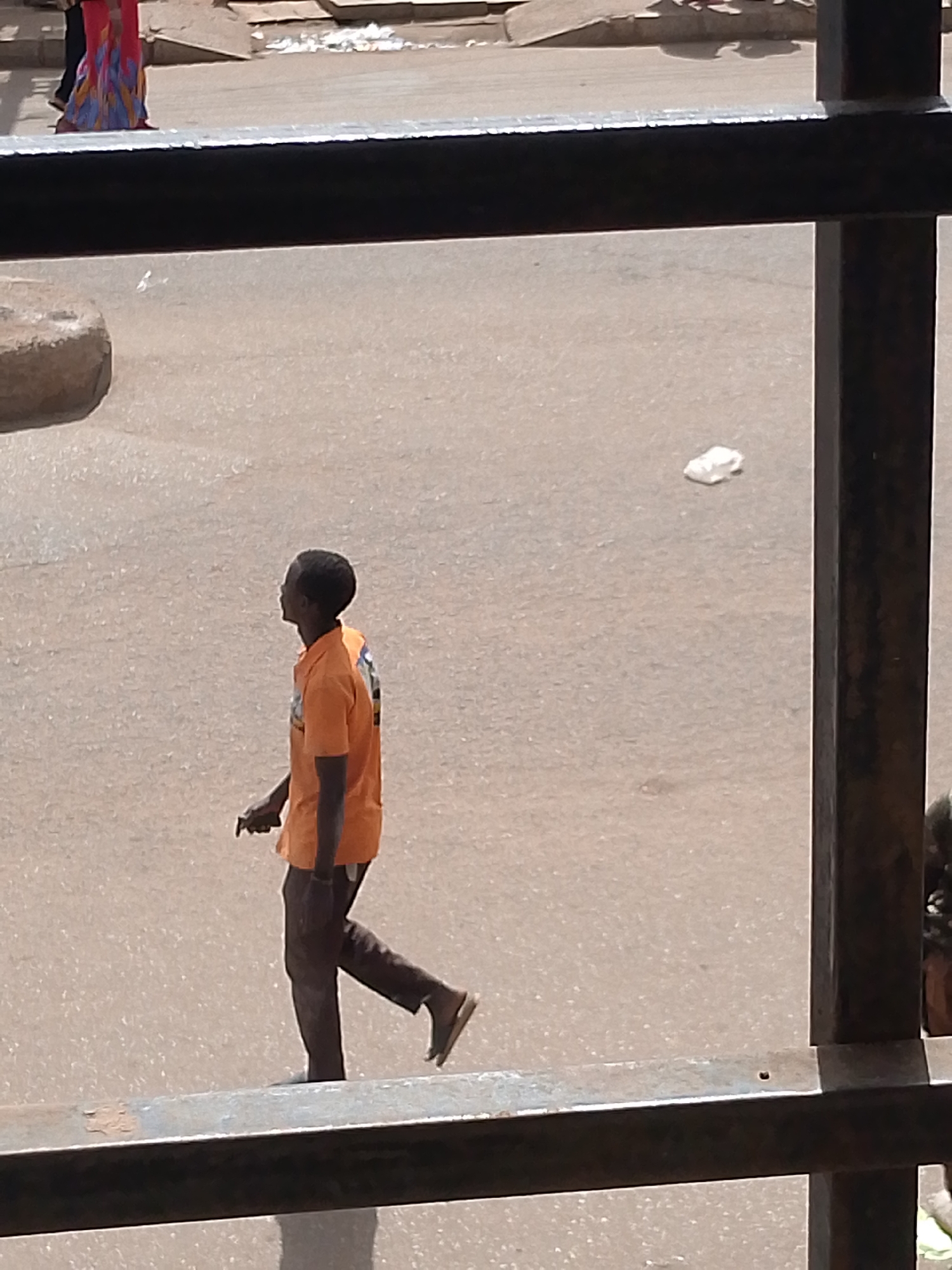 In Bukuru, Nigeria, a man in an orange t-shirt and dark pants walks across a paved street. He is viewed from an elevated perspective, through a dark metal railing. The man's back is to the camera, and he is looking to his right. In the background, a person in a colorful patterned dress is visible, as well as a white plastic bag on the street. The street appears to be a city street, with a curb visible on the left side of the image. The weather is sunny, as indicated by the shadows on the ground.