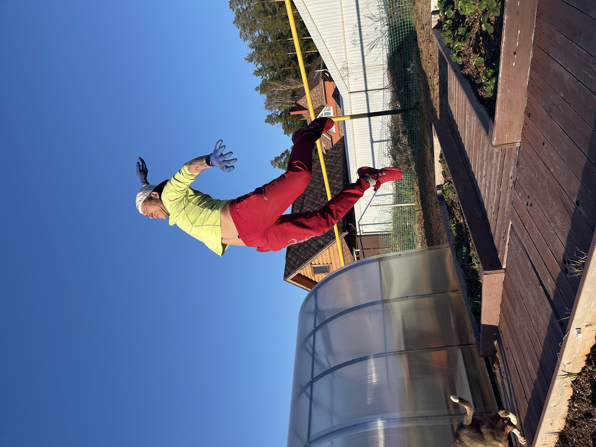 A man in a green shirt, red pants, and red shoes is airborne. He has a white head covering and blue gloves. The background shows a clear blue sky, a greenhouse, wooden decking, a fence, trees, and buildings. A dog is partially visible near the greenhouse. This image was captured in the city of Chaikovsky, Russia.