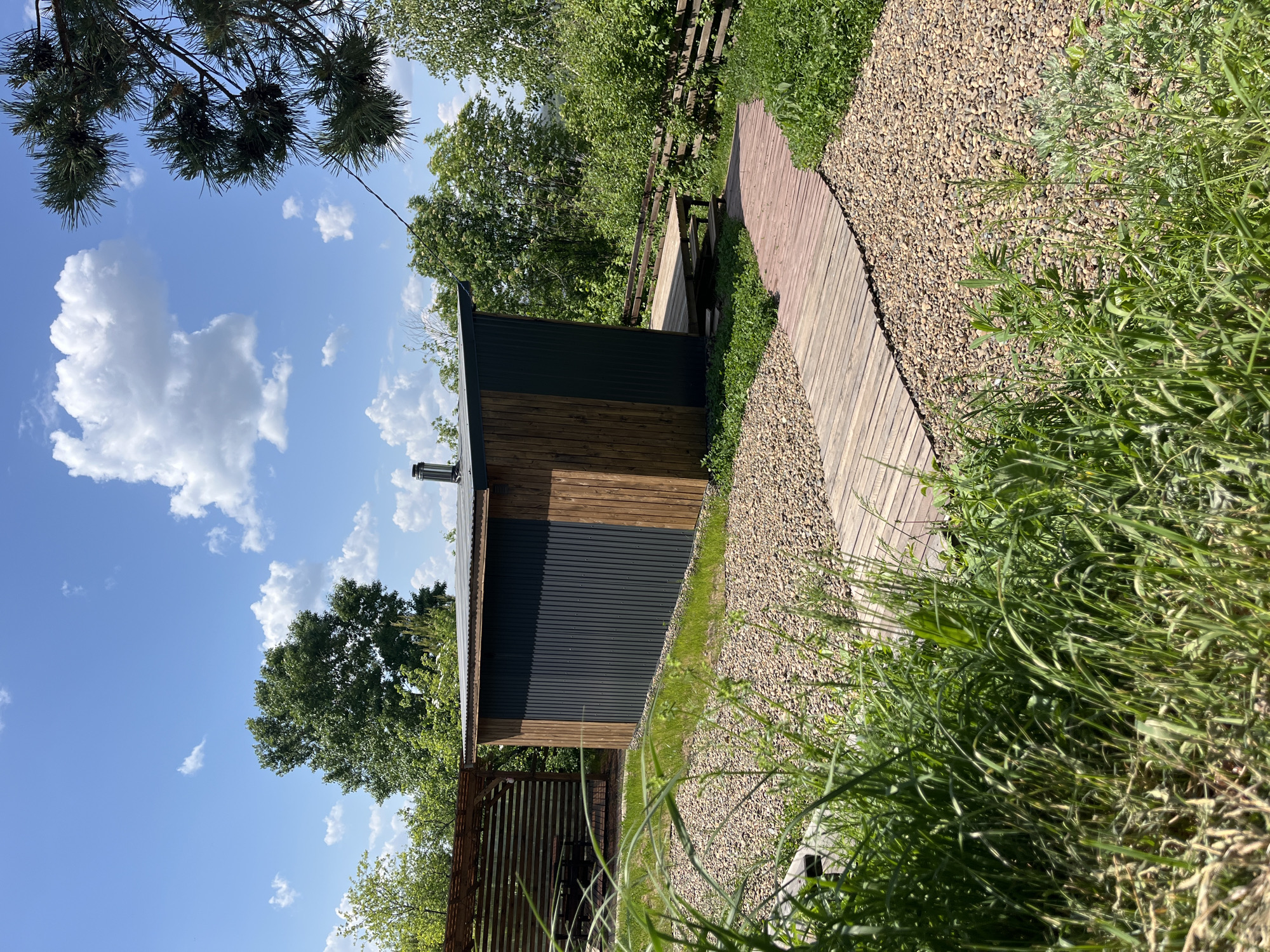 The image presents a scene in a likely rural or suburban setting near the city of Chaikovsky, Russia. A small building, possibly a sauna or shed, constructed with both wood and metal siding, stands to the left. The building has a chimney and is situated near a partially visible wooden pergola. A wooden walkway, bordered by gravel on one side and overgrown grass on the other, leads away from the building. The sky is visible with scattered clouds and a pine tree branch in the upper left corner. Other trees and vegetation surround the scene. There are no people in the frame.