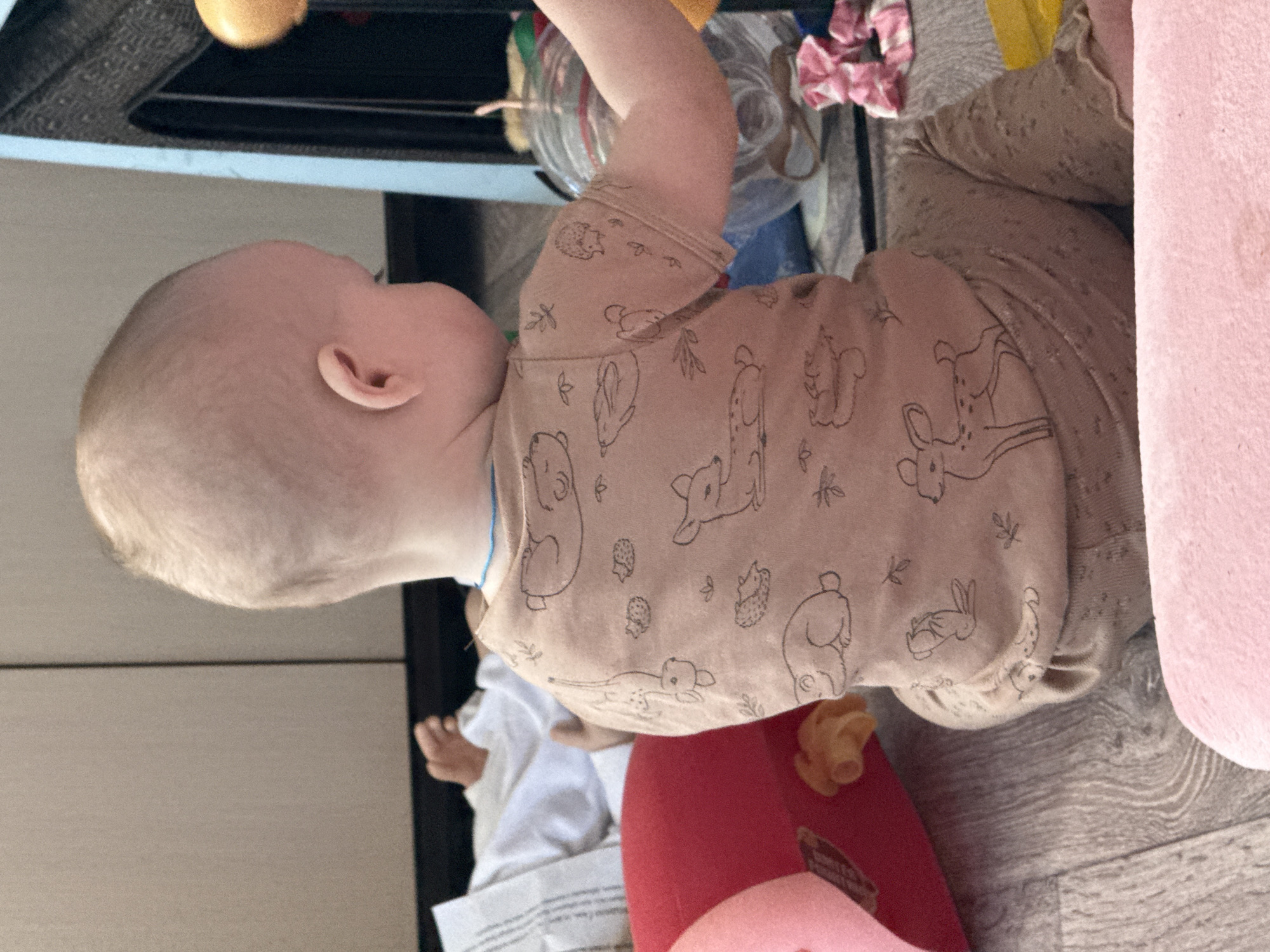 A baby lies on a floor next to a large, soft, pink stuffed animal, on a multi-colored, patterned surface. The baby is wearing a light brown shirt with cartoon animal designs and matching pants. The location is not indicated in the image.