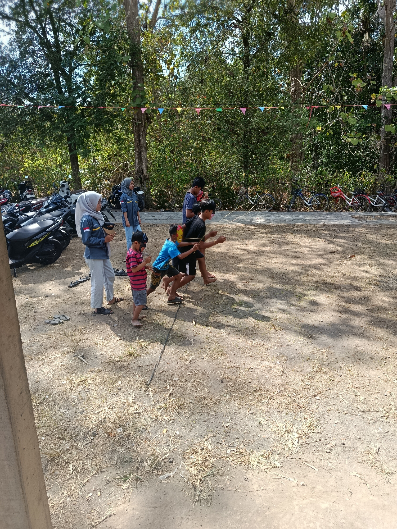 In Sedayu, Indonesia, at an outdoor event, several people are engaged in a game that appears to involve pulling a rope. In the foreground, a young boy in a striped shirt and grey shorts stands with a mask on his face, holding a rope. Beside him, another boy in a blue t-shirt and black shorts is also holding the rope. Behind them, a man in a black t-shirt and dark shorts is participating, his arm extended towards the rope. Further back, a young boy in a blue t-shirt and dark shorts is holding the rope, with another boy in a red and white striped shirt next to him. Two young women in matching blue shirts and grey pants are observing, one holding a phone. In the background, motorcycles are parked, and several bicycles are lined up. The ground is sandy with dry grass, and trees with green foliage surround the area, with a line of colorful triangular flags strung between them.