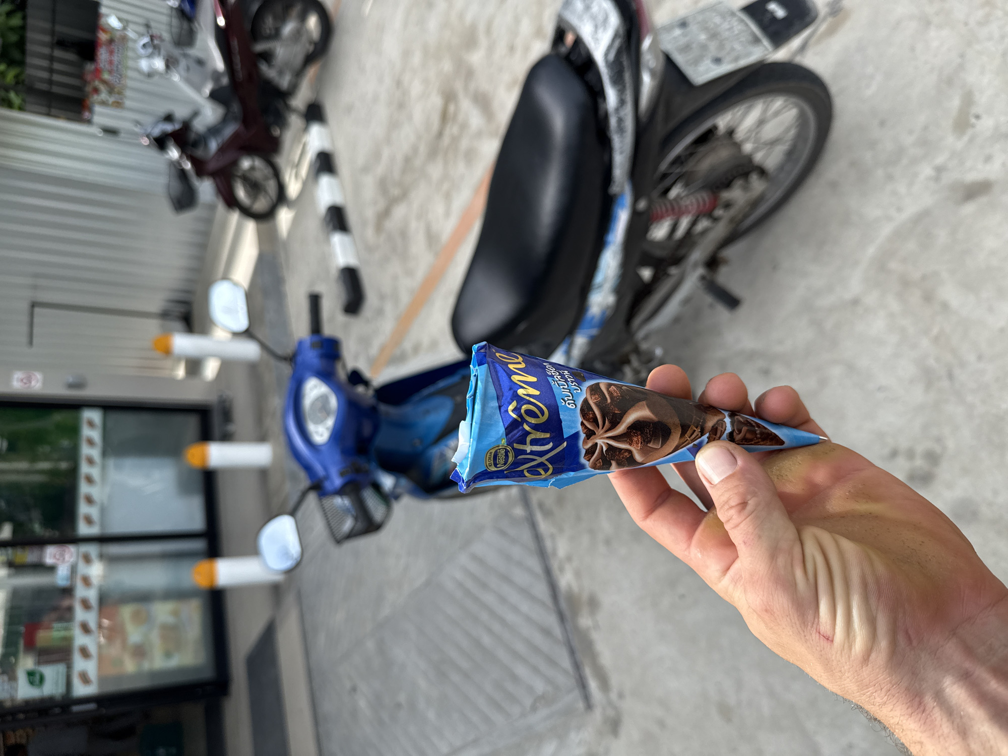 A first-person view shows a hand holding an "Extrême" chocolate ice cream cone in front of two parked motorcycles. The location is Baan Chaloklum, Thailand. In the background, a convenience store with visible product displays is discernible through the glass windows. The ground is concrete. The ice cream cone is partially unwrapped. One motorcycle is blue with orange tipped handle bars, the other is red.
