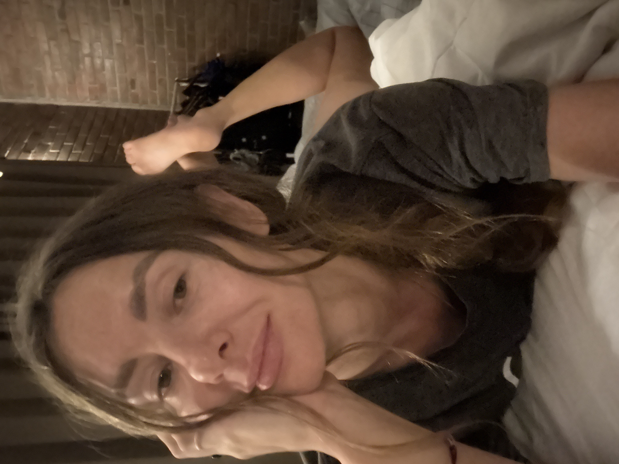 A woman with light brown hair is lying in a bed with white bedding. She is wearing a gray t-shirt and resting her face on her hand. Her legs are partially visible, extending towards the background. The background features a brick wall. The location appears to be in North Kuta, Indonesia.