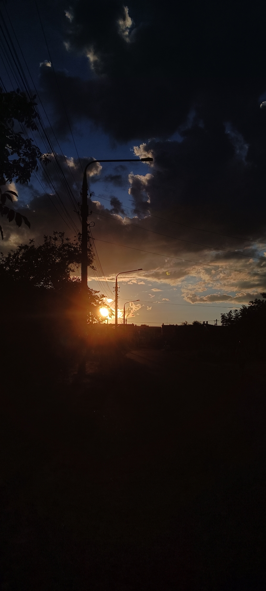 This is a vertical, low-angle shot of a sunset in Zaporizhzhia, Ukraine. The sun is low on the horizon, creating a bright lens flare that illuminates a section of the sky with warm orange and yellow hues. Dark, dramatic clouds dominate the upper portion of the frame, with lighter, wispy clouds scattered throughout. Silhouetted against the sunset are two streetlights on utility poles, and a line of trees and buildings along the horizon. The foreground is dark and indistinct, with the silhouette of trees and foliage on the left. The scene evokes a sense of dramatic beauty and the transition from day to night. The presence of utility poles and streetlights suggests an urban or suburban setting. The overall mood is somber yet hopeful, with the vibrant sunset offering a stark contrast to the encroaching darkness. There is no visible text in the image.
