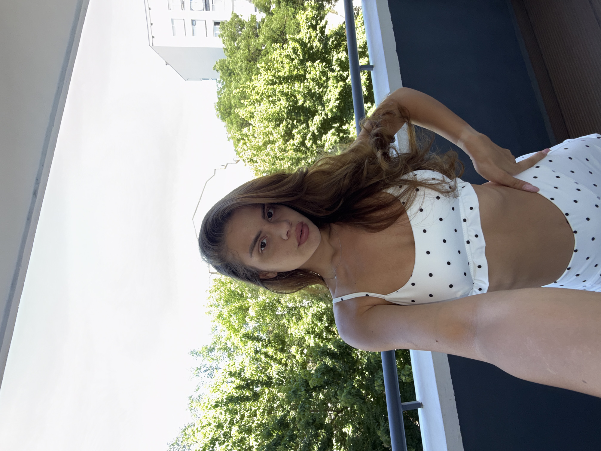 A young woman with long, wavy brown hair is pictured on a balcony. She is wearing a white polka dot top and white polka dot bottoms. Her left arm is bent at the elbow, with her hand resting on her hip. The background shows lush green trees and a tall white building, suggesting an urban setting with abundant foliage. The lighting indicates it is daytime, likely a sunny day. The woman's expression is neutral, with her gaze directed towards the camera. The image appears to be a casual, self-taken photograph.