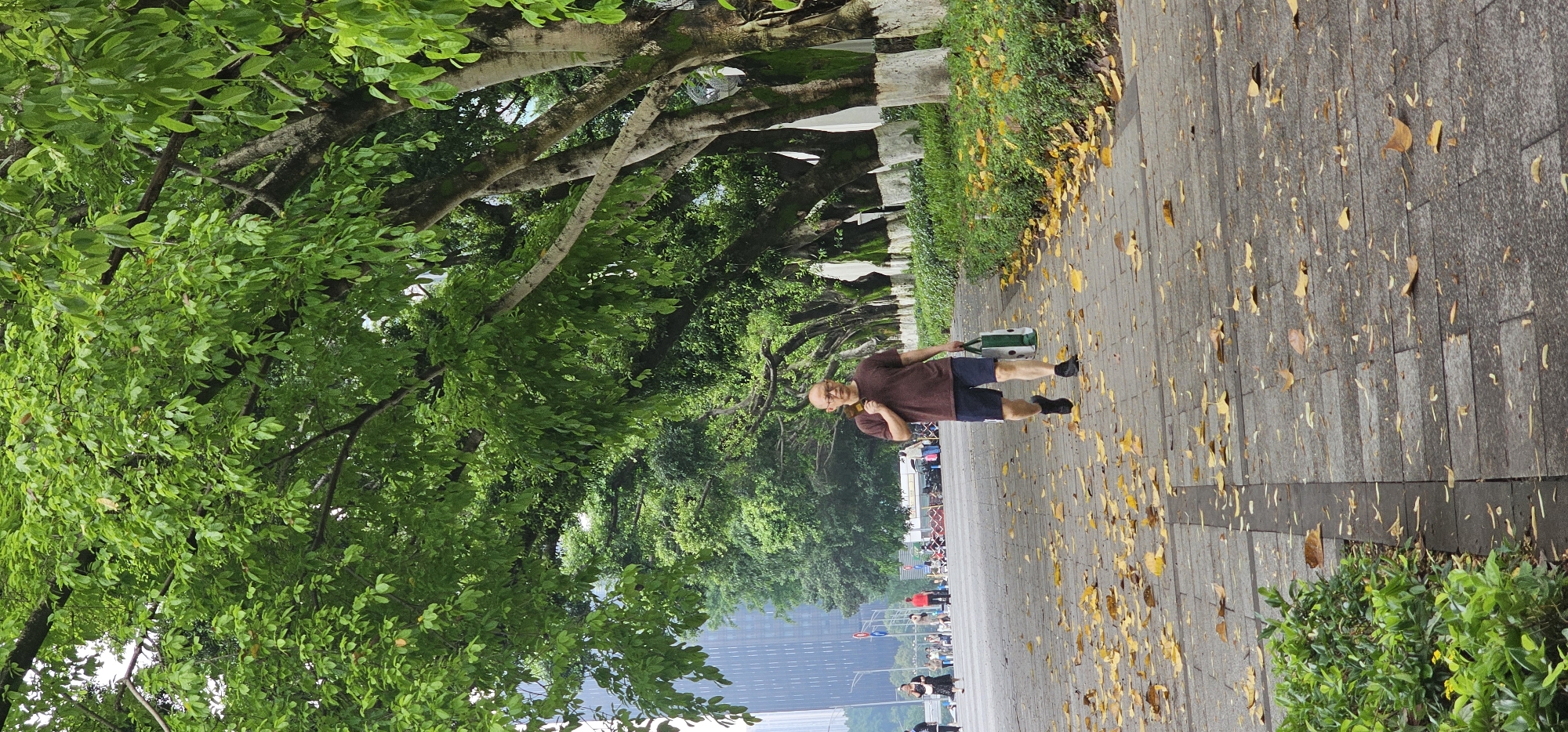 A horizontal, eye-level shot captures an outdoor scene in Jiulongpo District, China. A man walks to the right on a paved, leaf-strewn path lined with trees. He wears a brown shirt and blue shorts and carries a green and white spotted bag. Other people and vehicles are further down the path in the distance. Lush, green trees form a canopy above the walkway, casting shadows. Tall buildings can be seen in the background. The weather appears to be overcast.