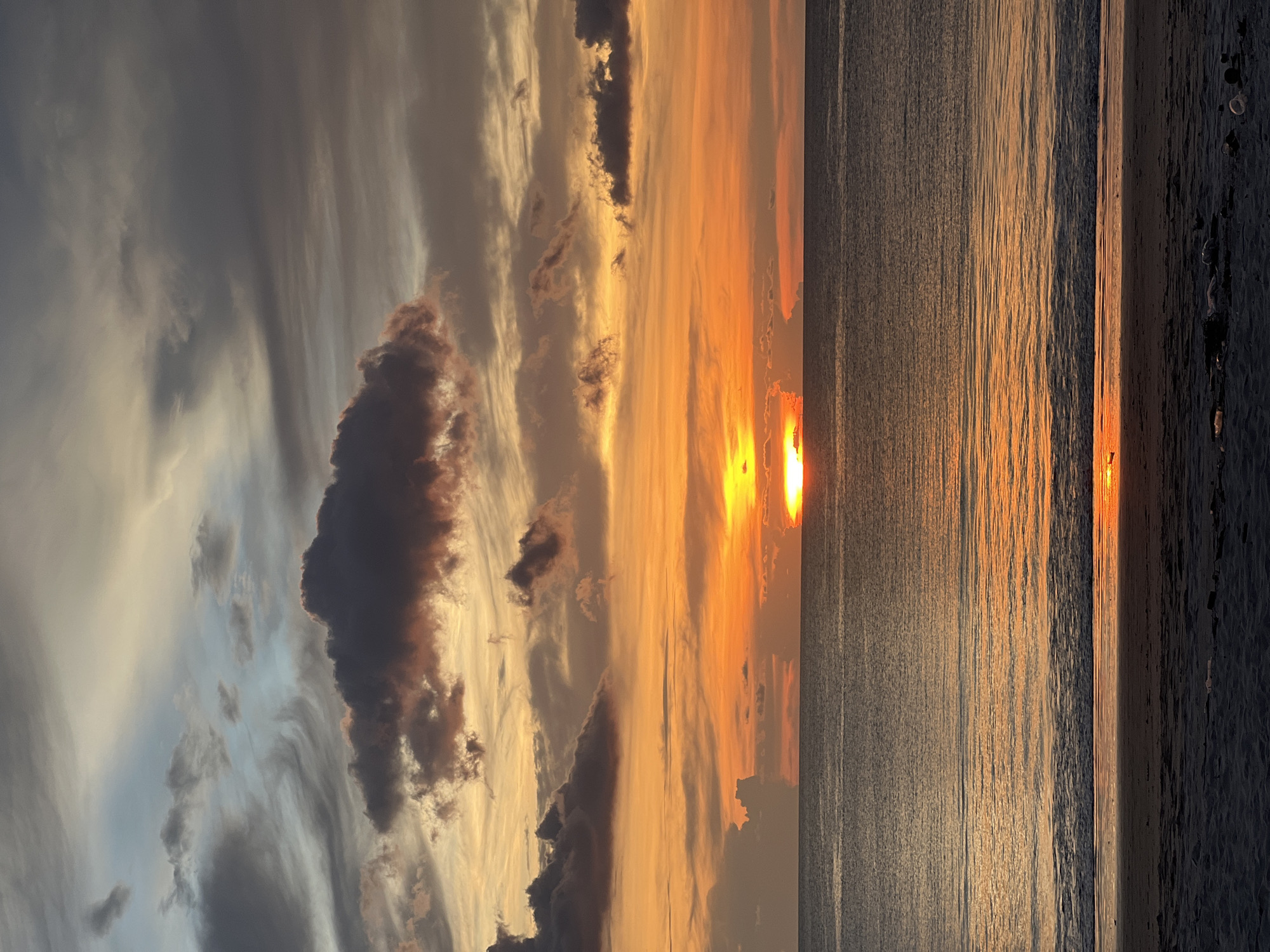 The image captures a sunset over a body of water, likely a beach. The sky is a mix of grey and orange hues, with various cloud formations including a large, dark cloud on the left side of the image. The sun is positioned near the horizon, reflecting its light onto the water's surface, creating a golden path towards the shore. The sand along the beach appears dark. The location is unknown.