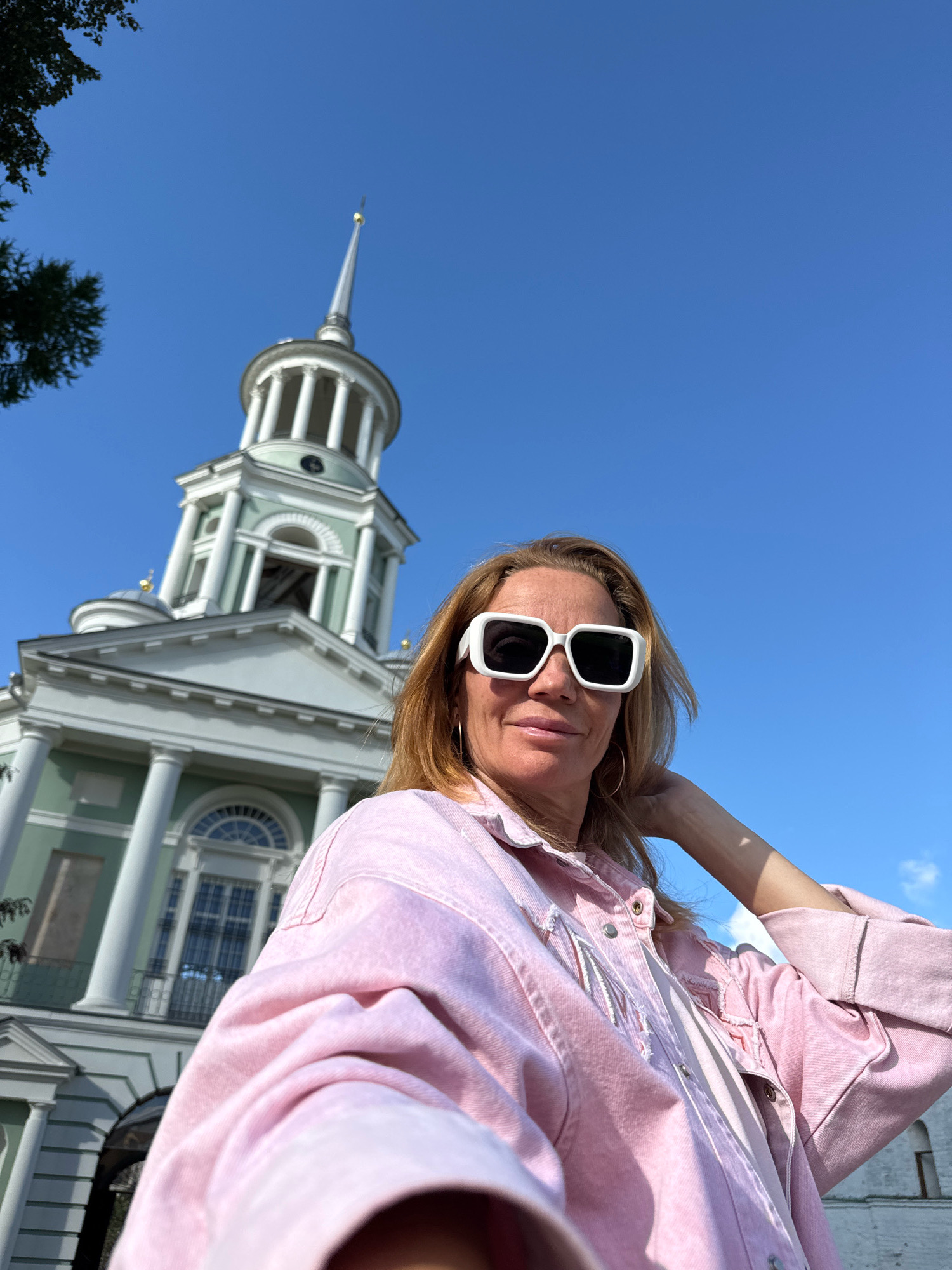 In the city of Torzhok, Russia, a woman with blonde hair and sunglasses poses for a selfie in front of the Borisoglebsky Monastery. She is wearing a light pink denim jacket and rests her hand on her head, looking towards the camera with a subtle smile. The monastery, a prominent architectural landmark, is visible behind her, featuring a white and green facade, classical columns, and a spire topped with a golden cross. The sky is clear and blue, indicating a sunny day.