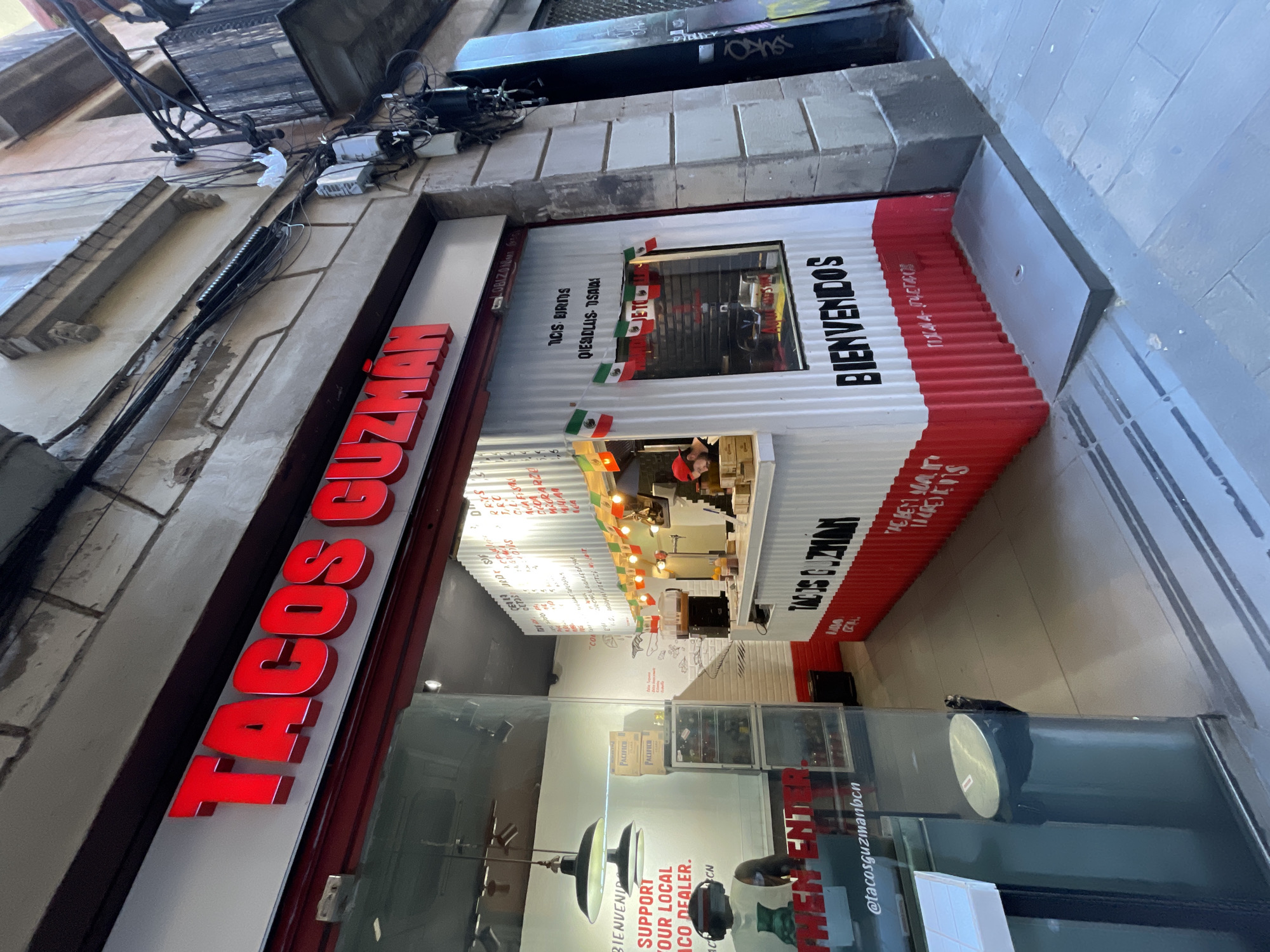 This media file captures the street-level exterior of a Mexican restaurant named "Tacos Guzman" in an urban setting. The storefront features a large, bright red sign with the words "TACOS GUZMAN" in prominent 3D letters. Below the sign, the facade mimics a shipping container, with alternating white and red corrugated panels.
On the white section of the facade, the word "BIENVENIDOS" is written vertically. Small Mexican flags are draped around a large window, through which the restaurant's interior is visible. Inside, a person in a red hat can be seen behind a service counter, illuminated by string lights. Other visible details inside include written text on the corrugated wall, potentially menu items, and stacked boxes labeled "PACIFICO."
The storefront window reflects elements from the interior and some external text, including "SUPPORT OUR LOCAL TACO DEALER" and an Instagram handle "@tacosguzmanbcn," which suggests its location in Barcelona. The building above the restaurant entrance shows typical urban architecture, with numerous electrical wires and cables attached to the exterior walls. The overall lighting suggests it is daytime, possibly overcast, or the indoor lighting creates a warm glow. The scene depicts a lively, informal eatery in a busy street environment.